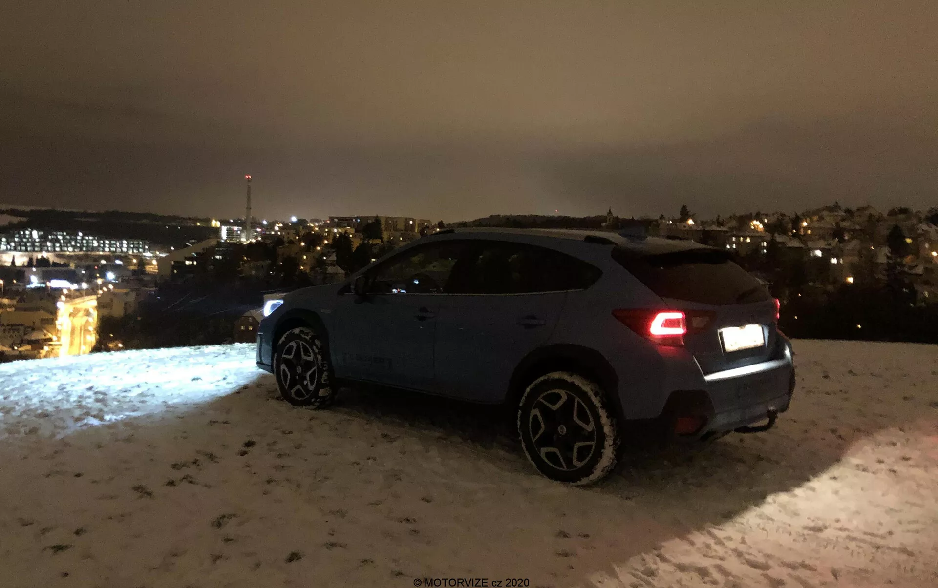 Subaru XV 1.6i Trend Lineartronic z 2018 roku sfotografowany nocą pod kątem trzech czwartych z tyłu, ukazujący prawą stronę i tył samochodu, w tym światła tylne, tylny zderzak, prawe tylne koło, drzwi pasażera i prawe przednie koło. Pojazd, prawdopodobnie w kolorze ciemnym niebieskim lub szarym, jest zaparkowany na powierzchni pokrytej śniegiem, z widocznymi w oddali światłami miasta. Tylne światła wskazują, że samochód może być włączony, a charakterystyczne większe koła wskazują na wyższy poziom wyposażenia lub sportową wersję. Scena podkreśla estetyczne i ambientowe ustawienie bez wyraźnej widoczności zaawansowanych funkcji technologicznych.