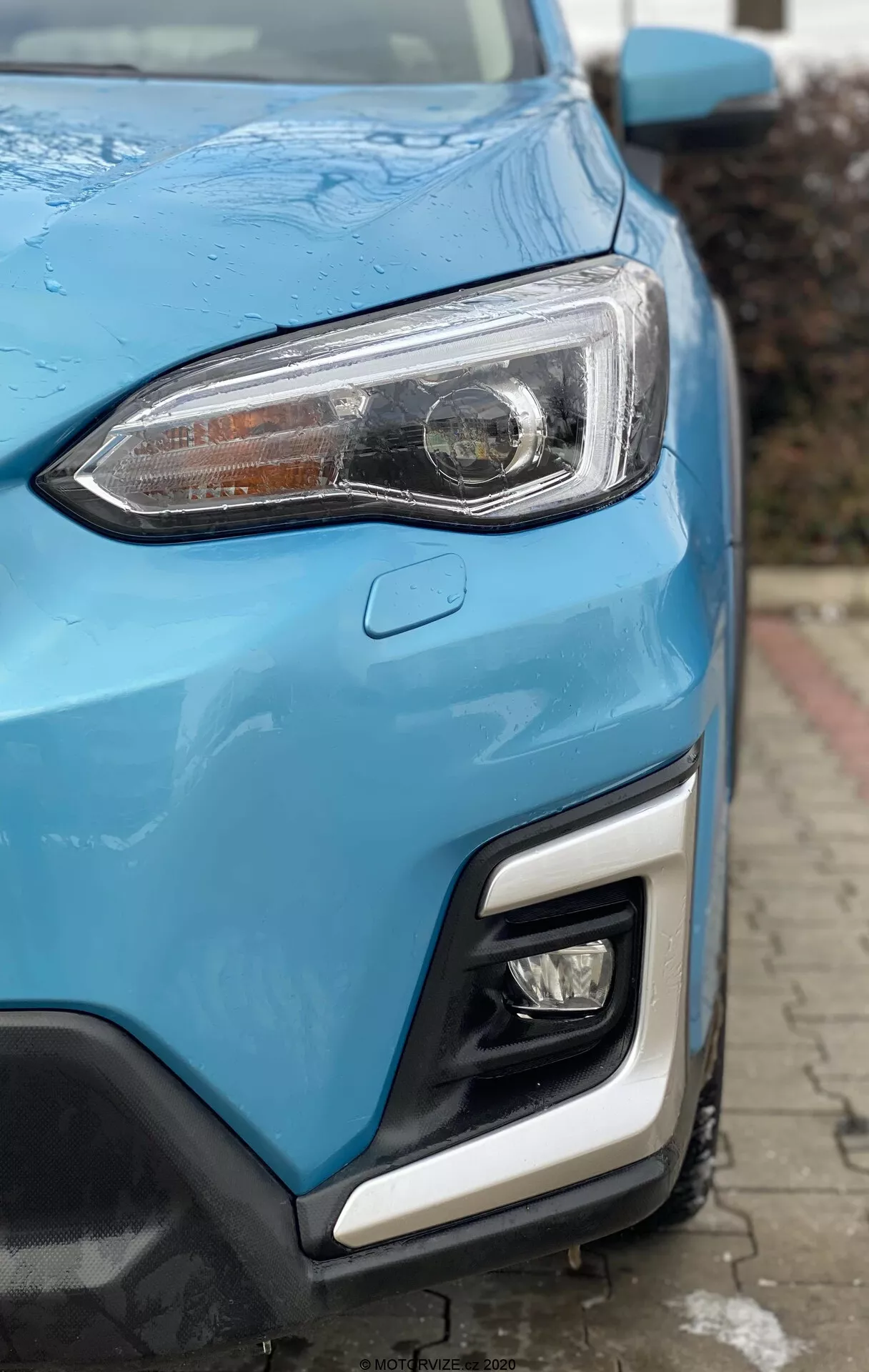 Zbliżenie na przedni róg Subaru XV 1.6i Trend Lineartronic z 2018 roku, pokazujące nowoczesny reflektor z przezroczystą soczewką i LED-owymi światłami do jazdy dziennej, część kratki i zderzaka w wibrującej niebieskiej farbie. Zderzak wyposażony w światło przeciwmgielne lub drógowe oraz czujnik parkowania. Eleganckie, sportowe panele nadwozia z kroplami wody sugerują niedawny deszcz lub mycie, podkreślając krzywiznę samochodu. Niewyraźne tło miejskie.