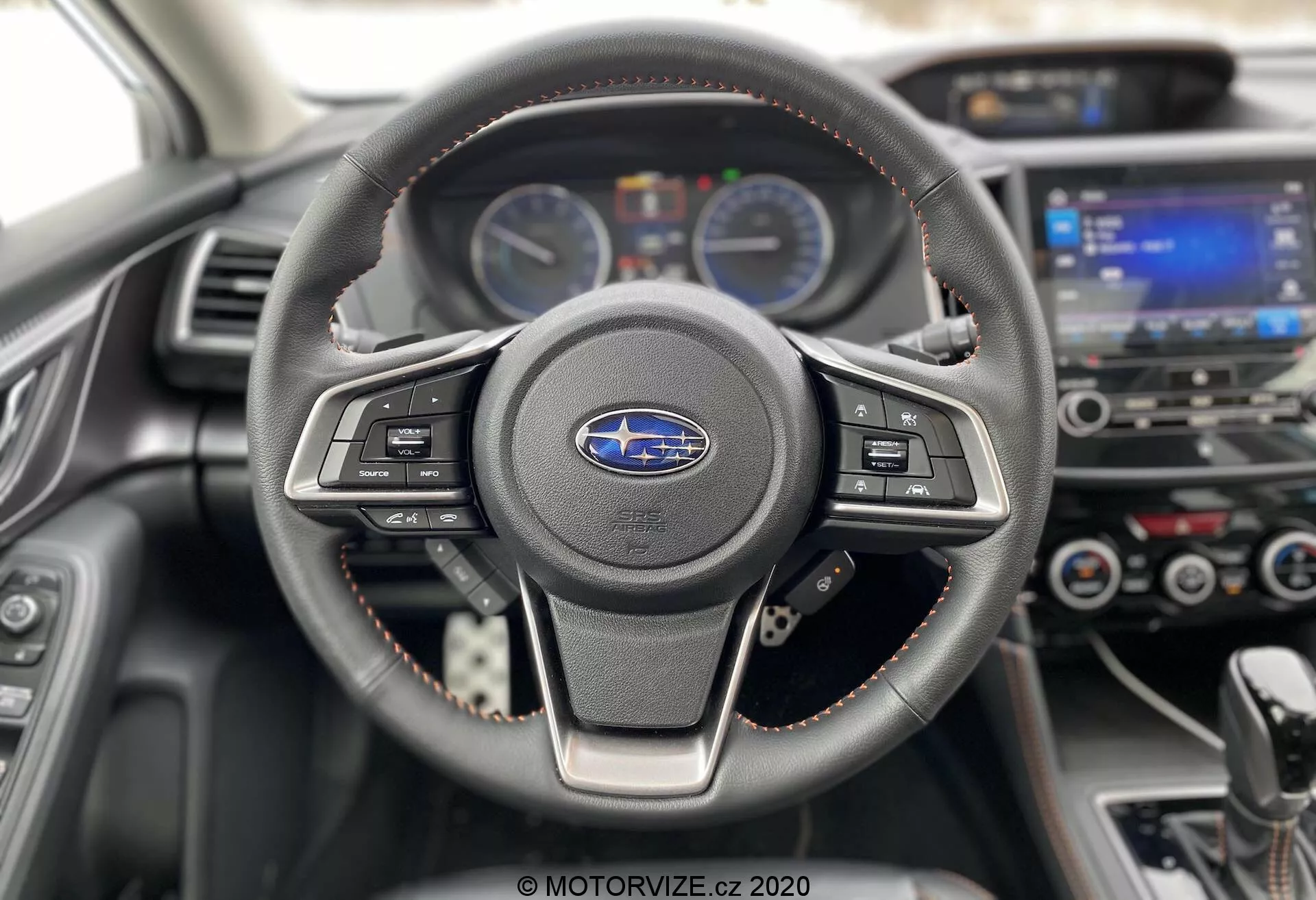 Wnętrze Subaru XV 2018 (Subaru-xv-1-6i-trend-lineartronic) skupiające się na kierownicy i desce rozdzielczej. Trójramienna, skórzana kierownica z widocznymi szwami posiada zamontowane przyciski multimedialne i tempomat. Za nią, deska rozdzielcza z dwoma analogowymi wskaźnikami, oddzielonymi cyfrowym wyświetlaczem informacji dla kierowcy. W tle, lekko rozmazany, widać dotykowy system infotainment oraz sterowanie klimatyzacją. Czerwone szwy na kierownicy i wyściółce deski rozdzielczej podkreślają sportowe lub premium wykończenie. Zdjęcie wykonane z perspektywy kierowcy, nowoczesne i dobrze utrzymane wnętrze, podkreślające technologię i design motoryzacyjny.