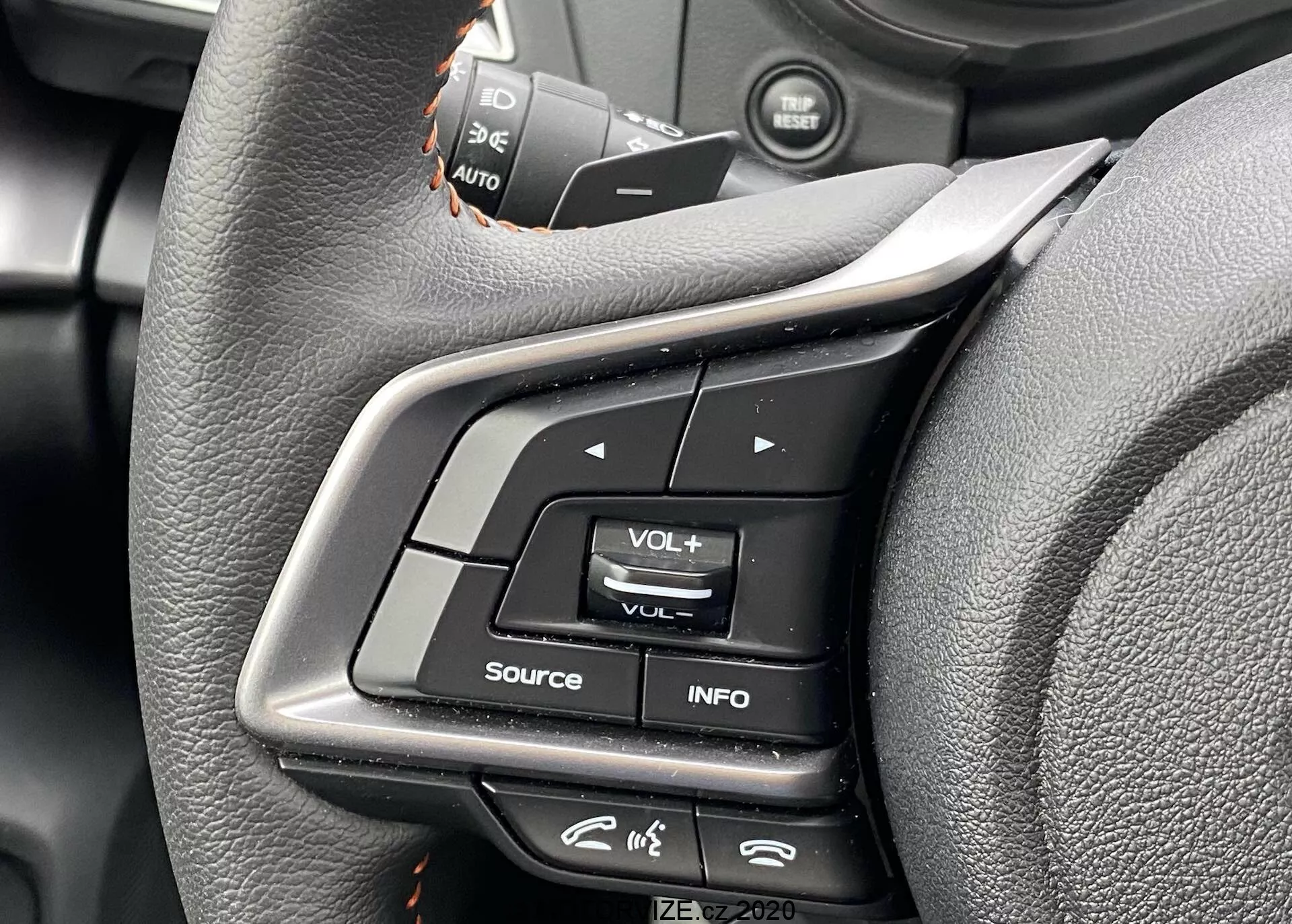 Zbliżenie na przyciski sterowania na kierownicy Subaru XV 2018 z perspektywy kierowcy. Zdjęcie podkreśla wielofunkcyjne przyciski na lewej stronie, w tym regulację głośności, przycisk INFO do ustawień wyświetlacza i ikonę przypominającą menu. Przyciski tempomatu są lekko rozmyte powyżej. Przyciski wykonane są z plastiku i gumy, z kierownicą owiniętą skórą o ergonomicznym kształcie. Metadane w prawym dolnym rogu wskazują 'IZE-EV2020'.