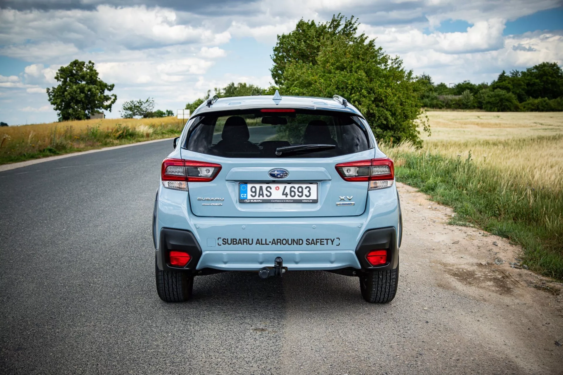 Tylny widok Subaru XV 2021 na wiejskiej drodze, pokazujący charakterystyczne tylne światła, podwójny układ wydechowy i klapę bagażnika z oznaczeniem bezpieczeństwa pod częściowo zachmurzonym niebem