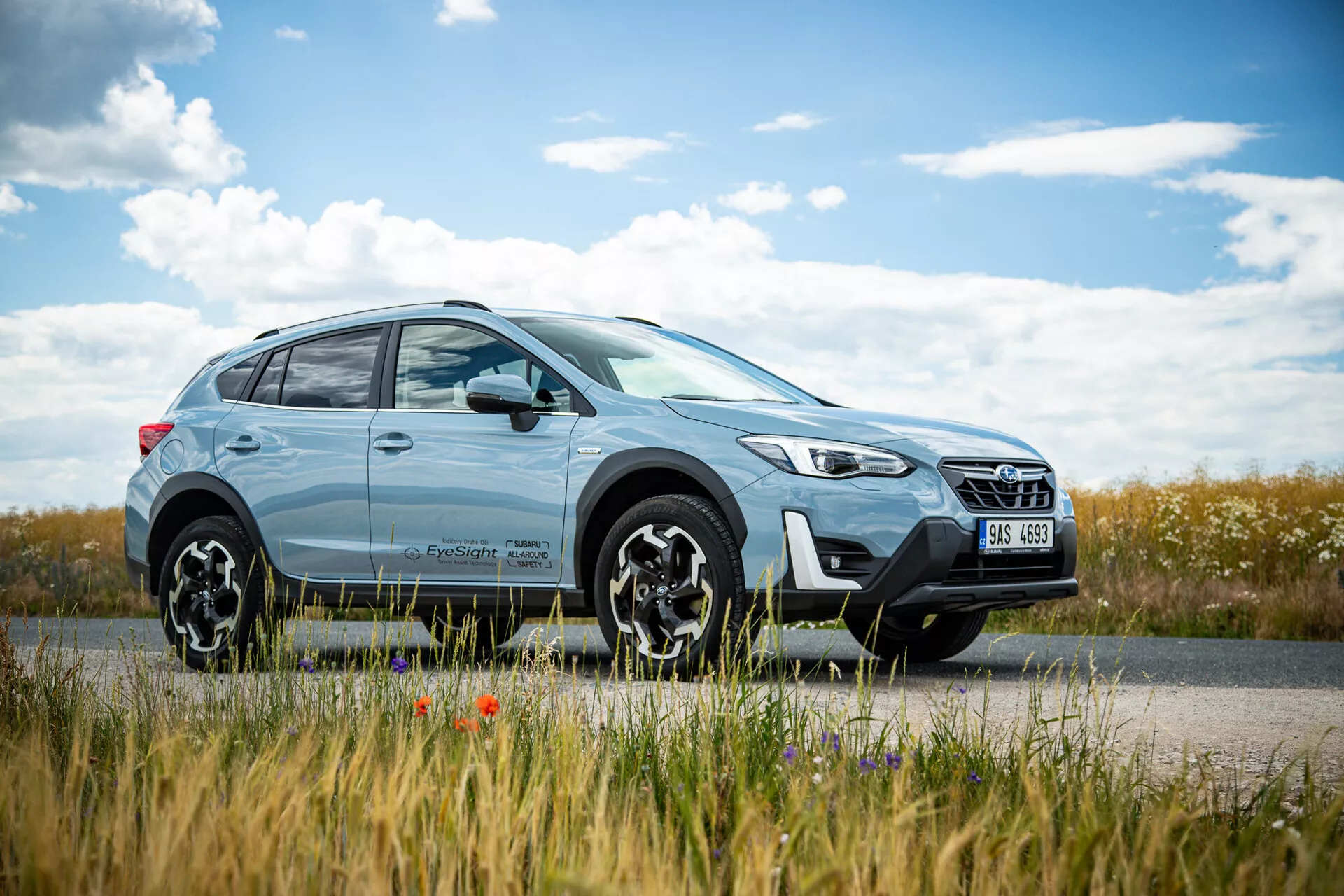 Jasnoniebieski crossover SUV Subaru XV 2021 zaparkowany przy drodze z krajobrazem wiejskim w tle, prezentujący nowoczesny design z reflektorami LED i kołami ze stopu wieloramienne, podkreślający zdolności zarówno do jazdy miejskiej, jak i terenowej.