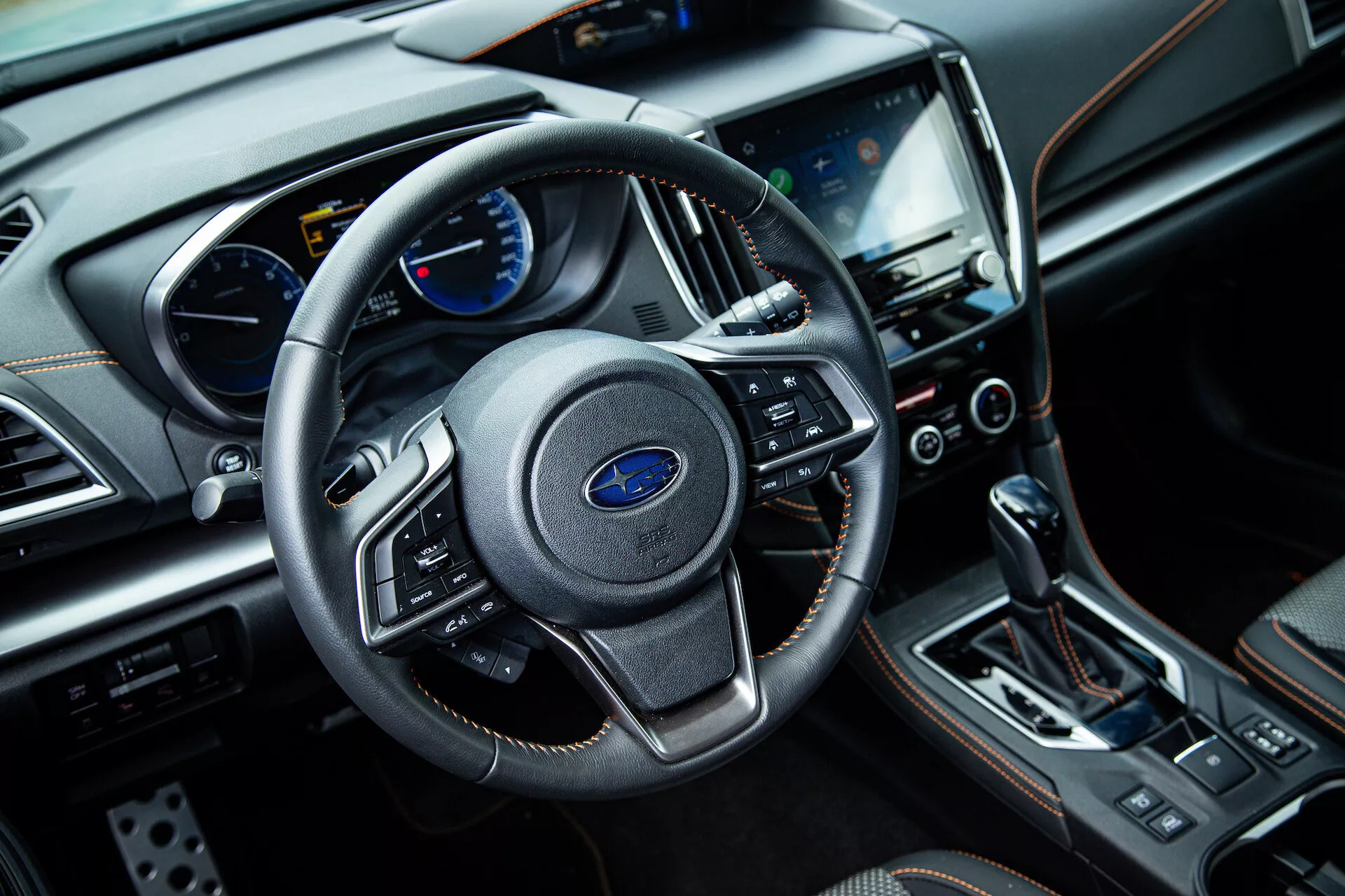 Widok wnętrza Subaru XV 2021 z perspektywy przedniego miejsca pasażera pokazujący trójramienne koło kierownicy z multifunkcyjnymi kontrolami, zespół wskaźników z analogowymi i cyfrowymi wyświetlaczami na desce rozdzielczej, centralny system infotainment z ekranem dotykowym, przyciski kontroli klimatyzacji, dźwignię automatycznej skrzyni biegów i luksusowe materiały wykończeniowe, w tym skórę i akcenty chromowane.