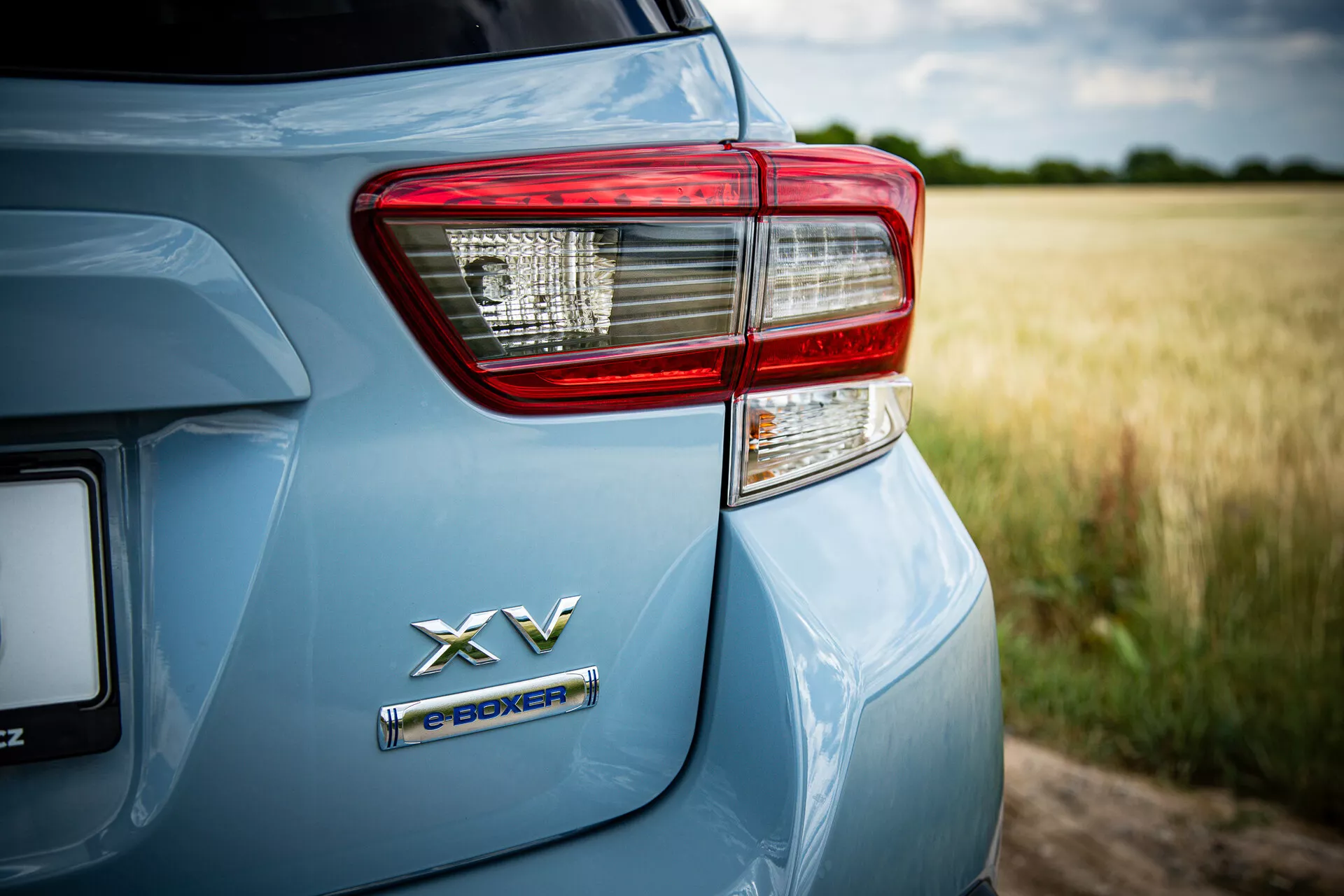 Zbliżenie na tylną lewą stronę Subaru XV 2021, uwypuklające emblemat napędu hybrydowego e-BOXER i szczegółowo opracowane zespoły świateł tylnych na metalicznym, jasnoniebieskim wykończeniu w wiejskim otoczeniu