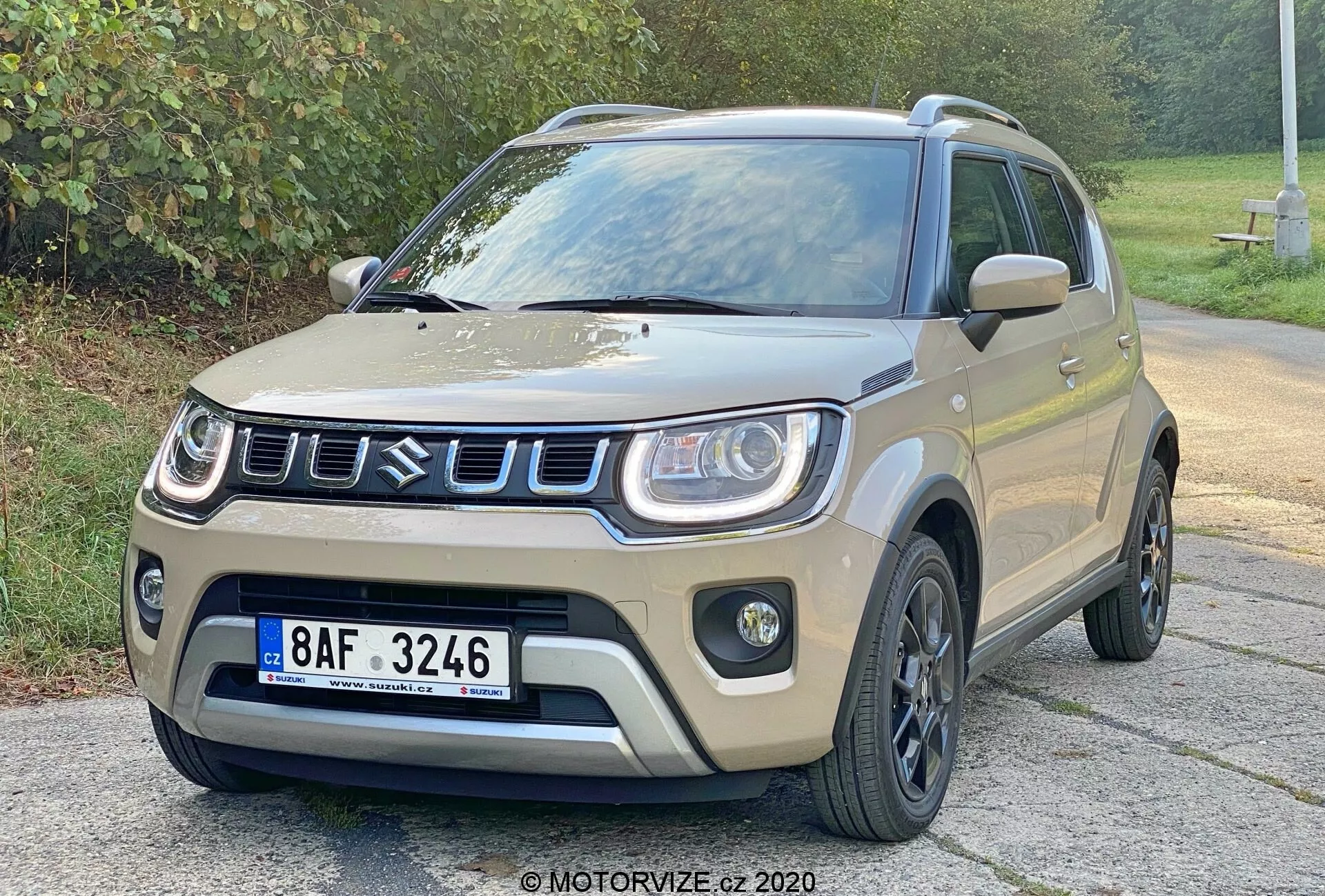 Kompaktowy SUV Suzuki Ignis 1.2 Dualjet Hybrid Club z 2020 roku, ukazany pod kątem trzech czwartych z przodu, prezentujący swoje jasnobeżowe lub piaskowe nadwozie z czarnymi akcentami na nadkolach, dolnych panelach bocznych i dachu. Przedni grill ma prostokątny wzór ze srebrnym lub chromowanym obramowaniem. Przedni zderzak ma widoczne obudowy świateł przeciwmgielnych oraz dynamiczną maskę z delikatnymi konturami. Czarne wieloramienne felgi aluminiowe oraz czarne lusterka boczne dodają solidnego wyglądu. Pojazd jest zaparkowany na asfaltowej powierzchni z zielonym listowiem w tle przy miękkim oświetleniu.