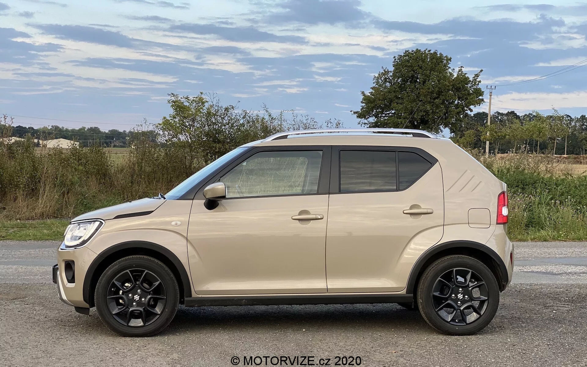 Profil boczny kompaktowego SUV-a Suzuki Ignis 1.2 Dualjet Hybrid Club 2020 z czterema drzwiami, czarnymi pięcioramiennymi alufelgami, dwukolorowym wystrojem w odcieniach beżu i czerni, przyciemnianymi szybami i możliwymi bocznymi lusterkami z wbudowanymi kierunkowskazami, w wiejskim otoczeniu.