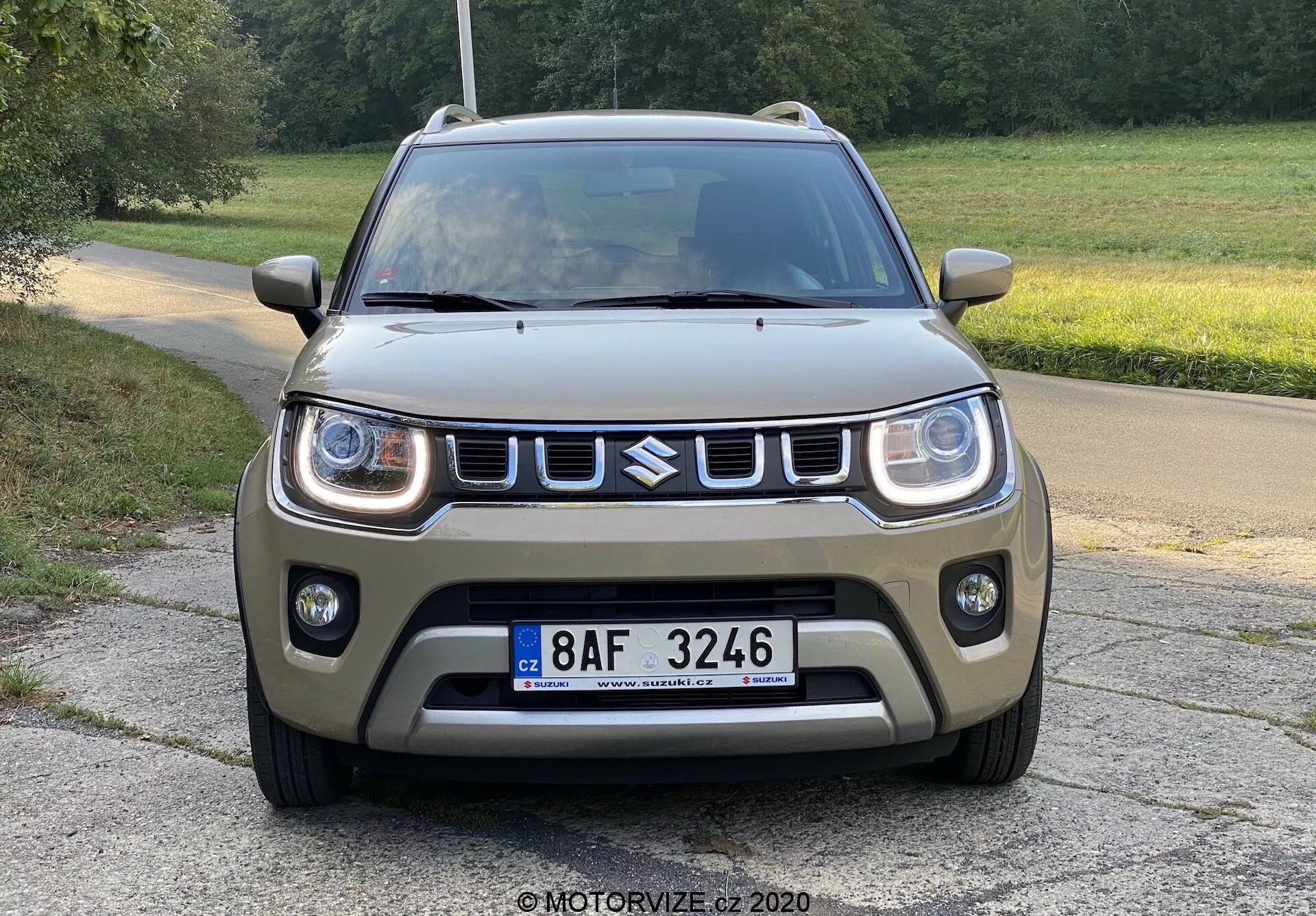 Widok z przodu kompaktowego SUV-a Suzuki Ignis 2020 na wybrukowanej drodze z trawiastym tłem, wiejskie lub parkowe otoczenie, zdjęcie z niskiego kąta podkreślające design przedniej części, z dziennymi światłami LED, wyraźnym grillem z oznaczeniem, masywnym przednim zderzakiem z światłami przeciwmgielnymi, matowe wykończenie w ziemistym zielono-brązowym kolorze