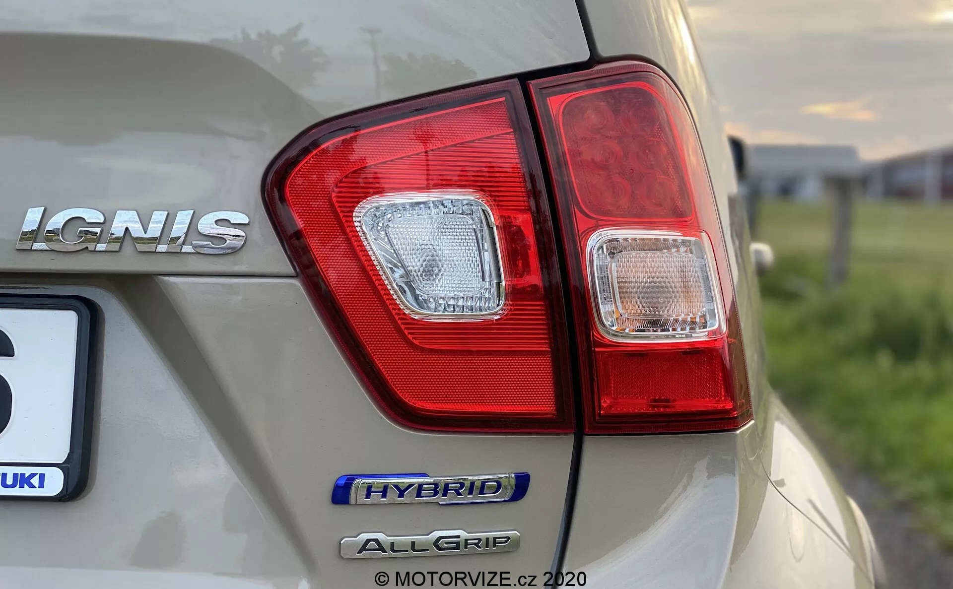 Zbliżenie tylnego narożnika Suzuki Ignis 2020, skupiające się na klastrze świateł tylnych i oznaczeniach modelu z niskiej, lekko nachylonej perspektywy. Na prawej stronie samochodu wyraźnie widoczne oznaczenia 'IGNIS', 'HYBRID' i 'ALLGRIP', podkreślające technologię hybrydową i system napędu na wszystkie koła. Samochód w jasnym, szampańskim kolorze ma błyszczące wykończenie z refleksami otoczenia, a obraz uchwyca wyraźne szczegóły grupy świateł tylnych z segmentami czerwonymi i przeźroczystymi, zachowując płytką głębię ostrości.