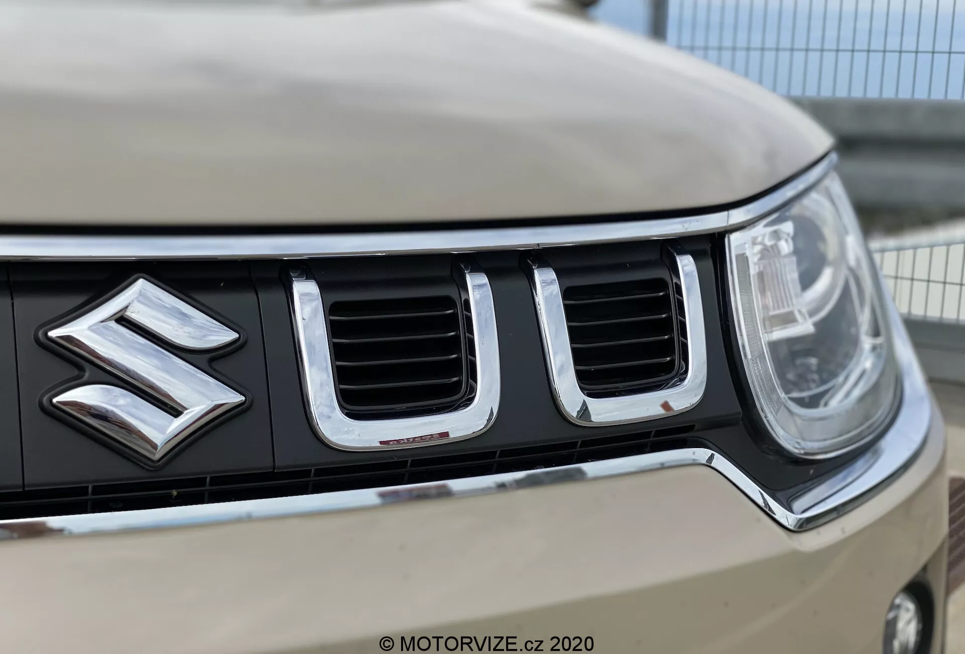 Zbliżenie na przednią kratkę oraz emblemat samochodu Suzuki Ignis 2020, podkreślające wyraźne logo Suzuki, pionowo zorientowane chromowane szczeliny, lewy reflektor z nowoczesną technologią LED lub HID oraz jasną karoserię. Płytka głębia ostrości podkreśla szczegóły kratki i logo, rozmywając jednocześnie dalekie krawędzie i tło.