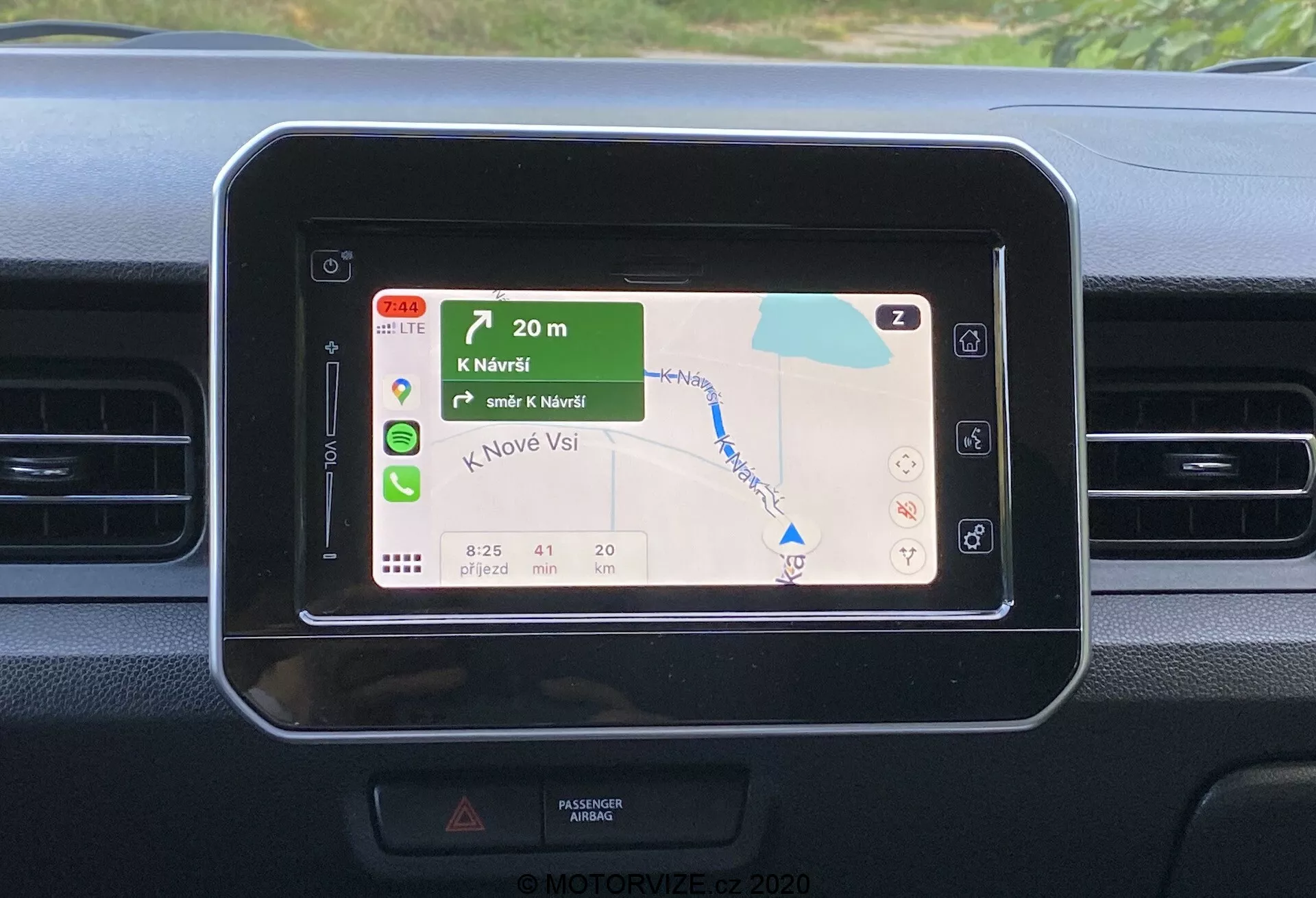 Zbliżenie deski rozdzielczej Suzuki Ignis 2020, koncentrujące się na prostokątnym ekranie systemu infotainment z zaokrąglonymi rogami, wyświetlającym kolorową mapę nawigacyjną GPS z zielonymi krajobrazami i niebieskimi zbiornikami wodnymi, pokazującym krok po kroku kierunek, ETA, odległość i limity prędkości. Deska rozdzielcza ma proste elementy projektowe z poziomymi wentylatorami w pobliżu, kontrastującymi materiałami i sugeruje obsługę dotykową lub zdalną bez widocznego kontekstu zewnętrznego.