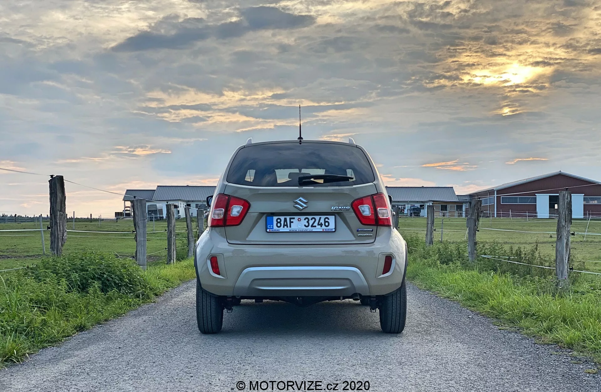 Widok z tyłu nieruchomego kompaktowego SUV-a Suzuki Ignis 2020 1.2 Dualjet Hybrid Club na wiejskiej drodze o wczesnym wieczorze, prezentującego nowoczesne tylne światła LED, tylny spojler, wysoko zamontowaną lampę stop, wycieraczkę, tylny zderzak z czujnikami parkowania oraz felgi aluminiowe, umieszczone na tle ciepłego zachodu słońca i otoczone drewnianymi ogrodzeniami oraz budynkami gospodarczymi.