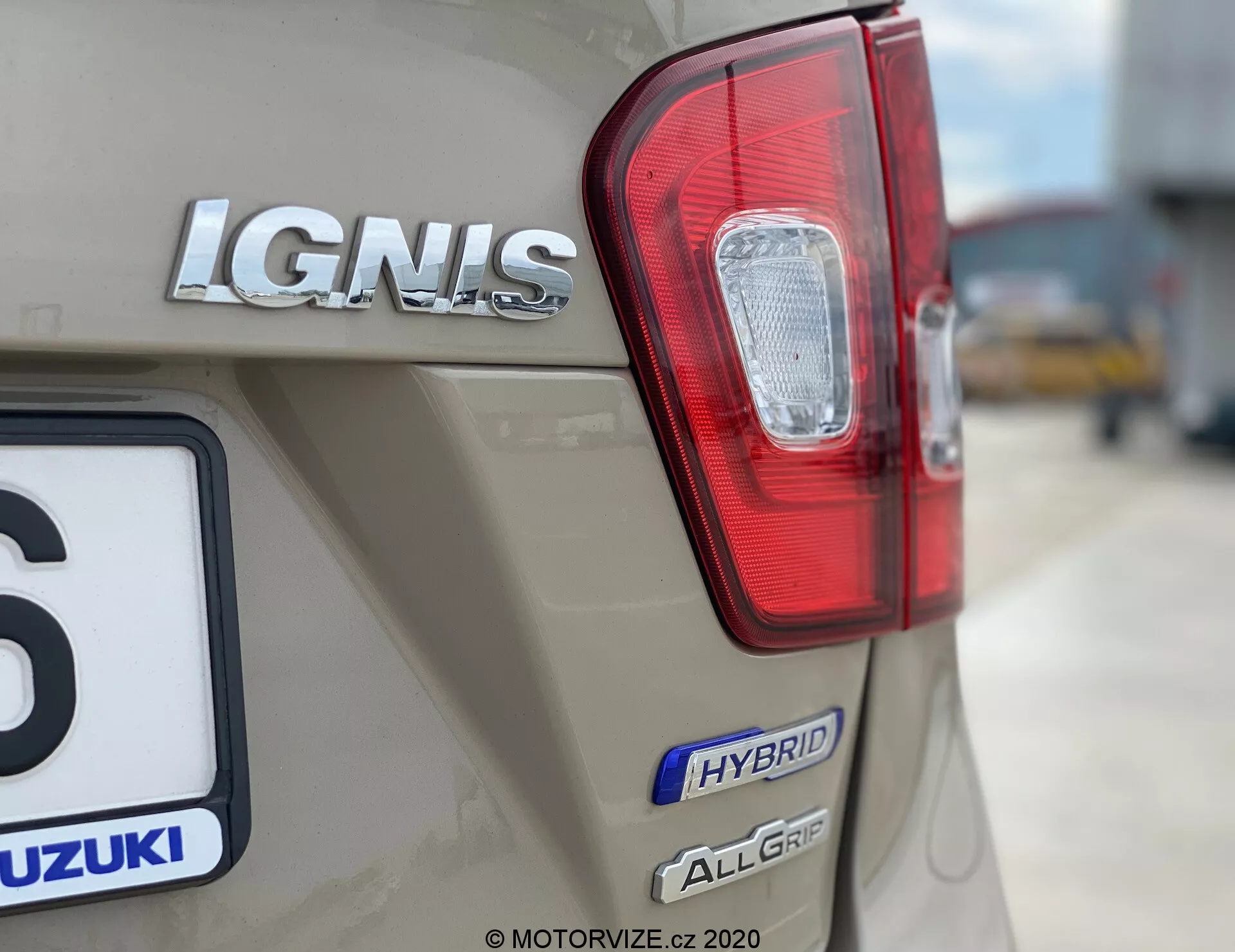 Zbliżenie tylnej części samochodu Suzuki Ignis 1.2 Dualjet Hybrid Club 2020, podkreślające prawe tylne światło z czerwonymi i przezroczystymi sekcjami, oznaczenie modelu 'IGNIS' w chromie, odznaka 'HYBRID' oraz odznaka 'ALLGRIP' wskazujące na technologię hybrydową i napęd na wszystkie koła. Logo Suzuki jest również częściowo widoczne nad tablicą rejestracyjną, a samochód ma metaliczne wykończenie w beżowym lub jasnobrązowym odcieniu. Tło jest rozmyte, aby podkreślić branding i cechy technologiczne pojazdu.