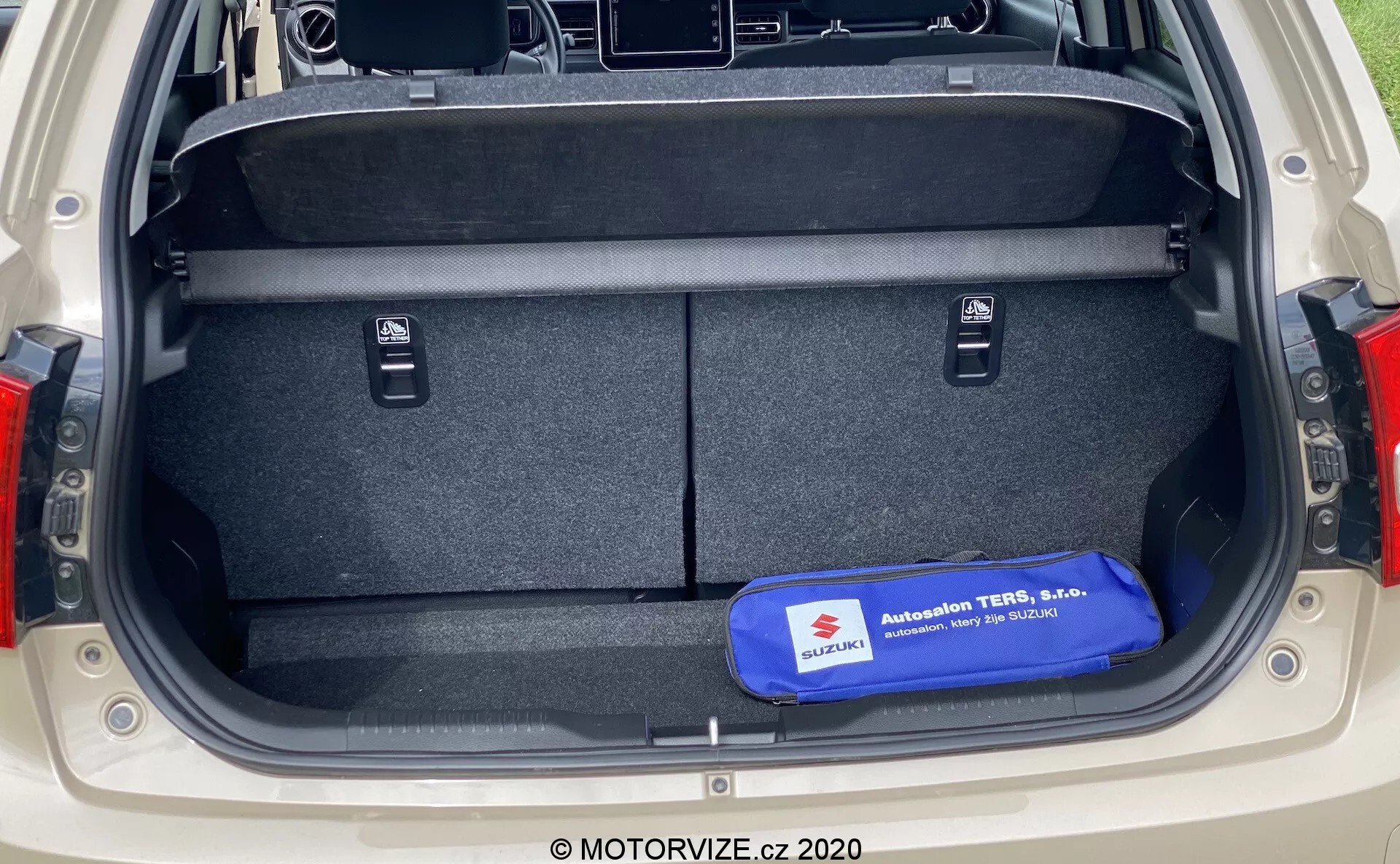 Bagażnik Suzuki Ignis 2020 widziany z perspektywy tylnej, ukazujący duże prostokątne miejsce do przechowywania z wykładziną na podłodze i bokach, małe wnęki na przedmioty, uchwyty do składania tylnej kanapy, niebieski zestaw bezpieczeństwa lub narzędzi na podłodze, nowoczesny design tylnych świateł widoczny po bokach bagażnika.