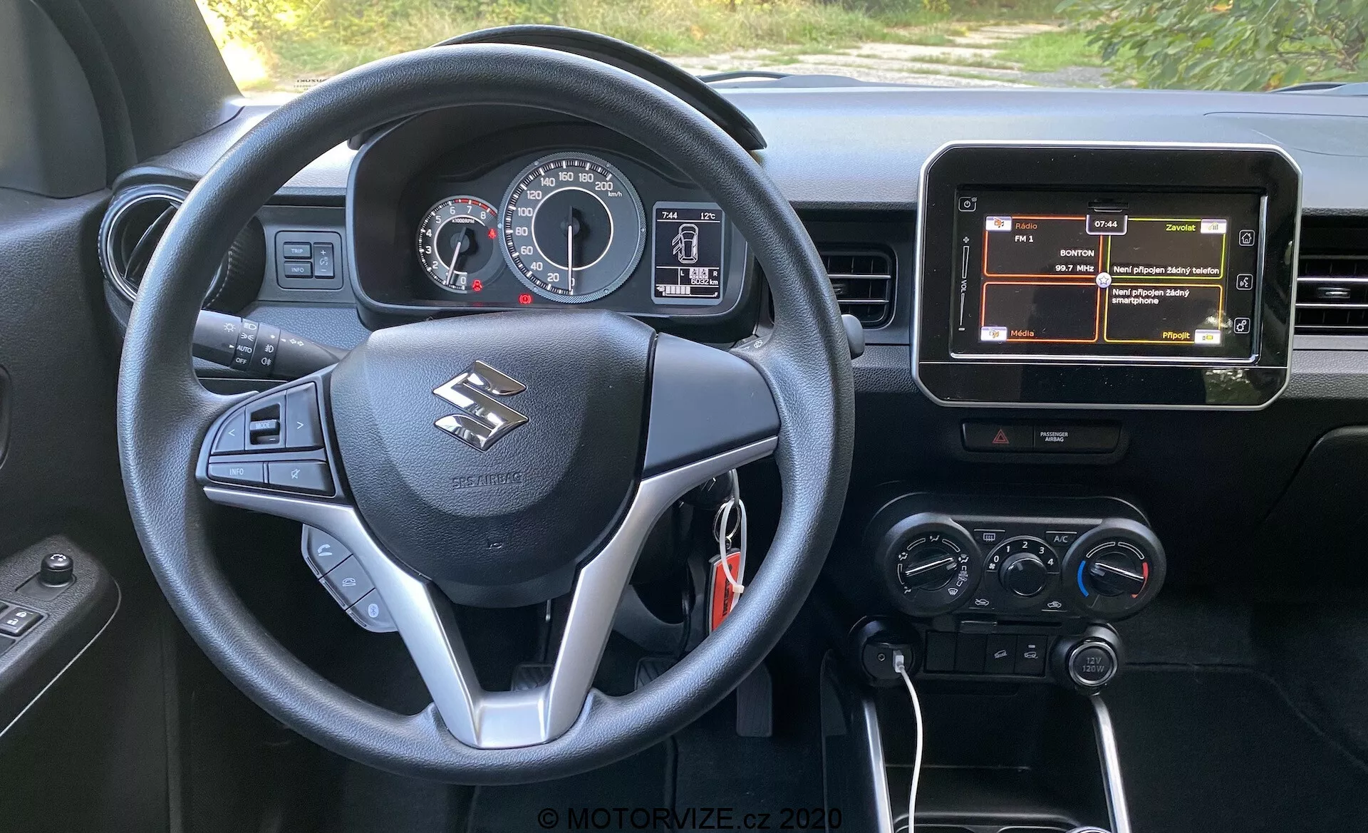 Widok wnętrza z perspektywy kierowcy Suzuki Ignis 1.2 Dualjet Hybrid Club 2020, prezentujący wielofunkcyjną kierownicę z przyciskami do sterowania dźwiękiem, telefonem i informacjami o pojeździe, zestaw wskaźników z analogowymi miernikami i centralnym wyświetlaczem cyfrowym, centralny ekran infotainmentowy z nawigacją oraz pokrętła kontroli klimatu. Pojazd ma automatyczną skrzynię biegów wskazywaną przez 'P' na dźwigni zmiany biegów, co sugeruje, że jest zaparkowany, oraz czarne wnętrze ze srebrnymi akcentami.