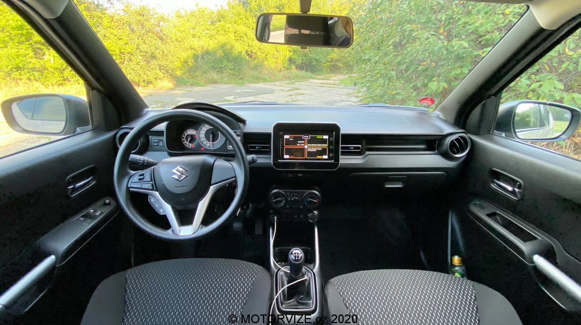 Widok wnętrza Suzuki Ignis 2020 z tylnego siedzenia pokazujący deskę rozdzielczą, kierownicę, system infotainment i zestaw wskaźników. Konsola środkowa z manualną skrzynią biegów, czarne wnętrze z różnorodnymi teksturami i materiałami.