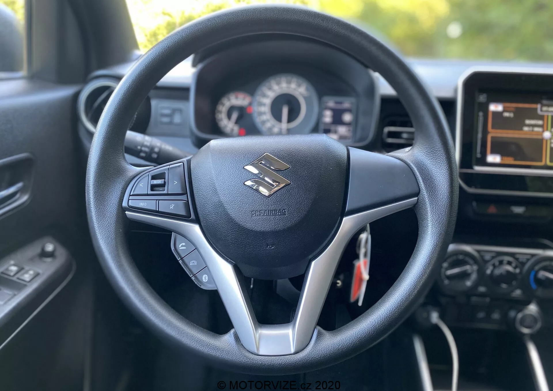 Widok wnętrza Suzuki Ignis 2020 z perspektywy kierowcy, ukazujący centralnie umieszczoną kierownicę o trzyramiennej konstrukcji ze srebrnym akcentem. Kierownica ma zamontowane przyciski do sterowania audio i prawdopodobnie tempomatem. Za nią widoczny jest zestaw wskaźników z prędkościomierzem i obrotomierzem oraz małym cyfrowym wyświetlaczem. Obraz uchwyca również część konsoli środkowej, w tym dotykowy system infotainment i dodatkowe przyciski. Wnętrze wyraźnie projektuje nowoczesny design z czarną kolorystyką, syntetyczną skórą i plastikowymi elementami, podkreślającymi wygodę kierowcy i technologię.