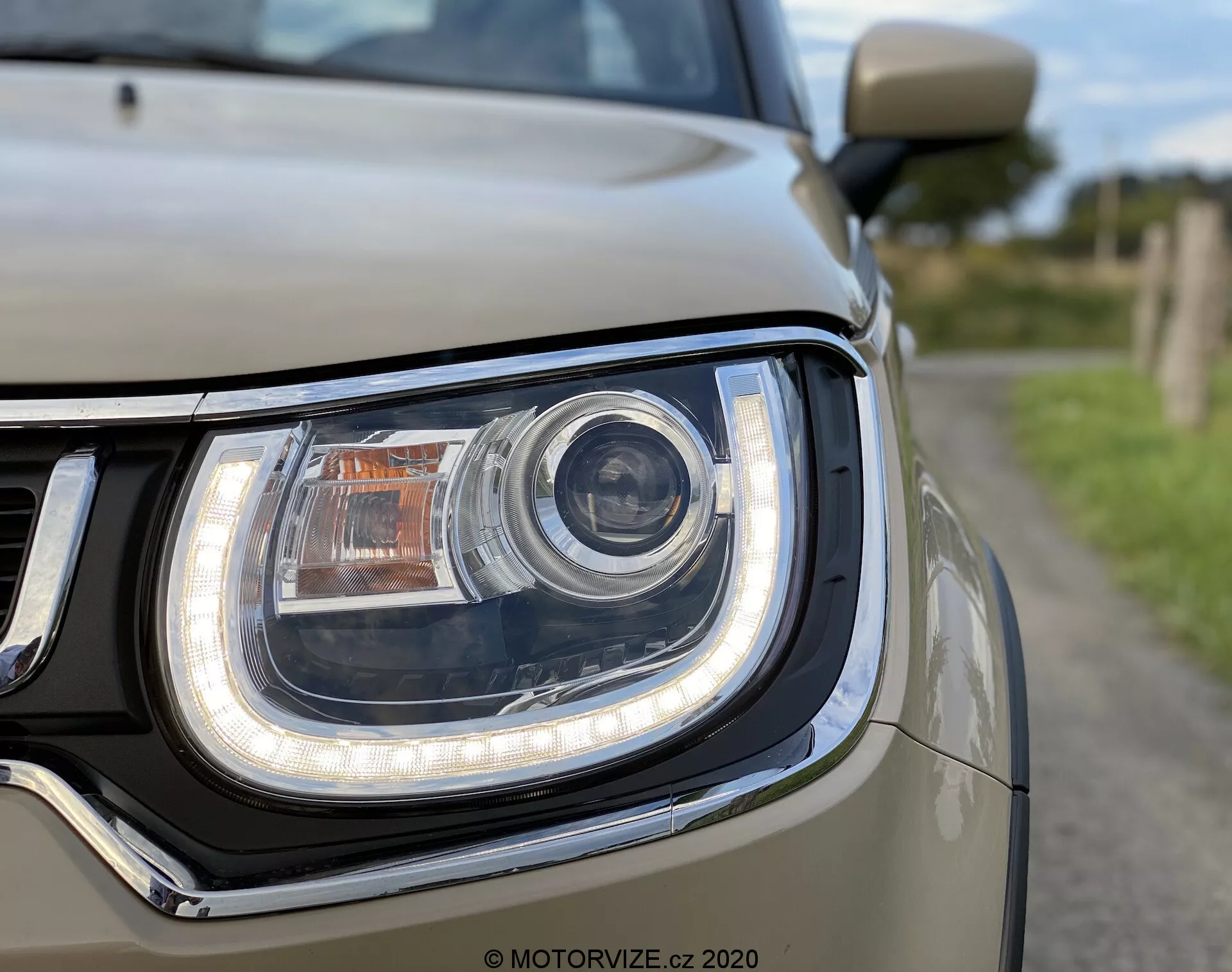 Zbliżenie jednostki przedniego lewego reflektora w Suzuki Ignis 2020, z centralnie umieszczonym głównym wiązką w stylu projektora, diodami LED obrysowującymi zakrzywiony kształt reflektora, pomarańczowym kierunkowskazem na górze oraz jasnokolorowym nadwoziem, prawdopodobnie srebrnym lub białym, otaczającym reflektor. Po lewej stronie widoczna jest sekcja chromowanej kratki, z płytką głębią ostrości podkreślającą skomplikowany design i technologię oświetlenia.