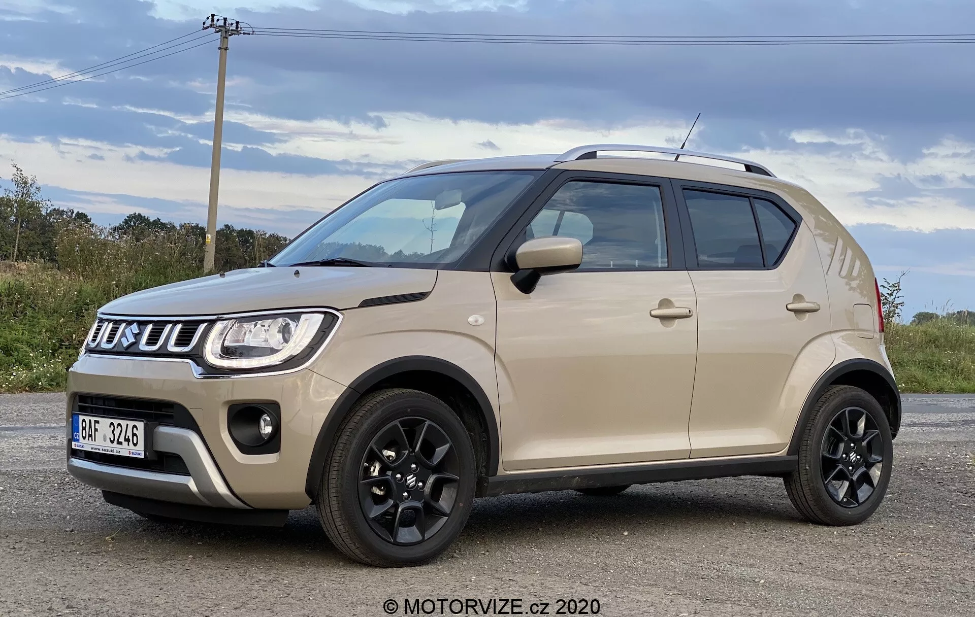 Widok z przodu w trzech czwartych na Suzuki Ignis 1.2 Dualjet Hybrid Club 2020, prezentujący jego współczesny, kompaktowy design SUV z dwukolorowym nadwoziem, wyrazistą kratką, reflektorami i solidnymi kołami. Nowoczesną estetykę pojazdu podkreśla czarny dach, lusterka, listwy boczne oraz duże opony o niskim profilu, co wszystko wskazuje na wysokie prześwit, odpowiednie do różnych warunków jazdy.