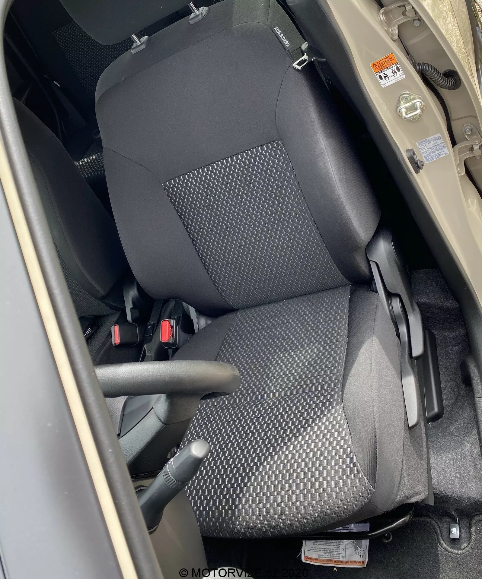 Zbliżony widok z góry przedniego siedzenia pasażera w Suzuki Ignis 2020, pokazujący zagłówek, oparcie i część siedząca. Zdjęcie wykonane z perspektywy osoby stojącej obok otwartych drzwi znajdujących się wokół słupka B. Siedzenie tapicerowane jest czarną tkaniną z teksturowanym wzorem w kropki i posiada ręczną regulację. Widoczny jest trzypunktowy pas bezpieczeństwa, a beżowe lub jasnobrązowe wnętrze kontrastuje z czarnym siedzeniem.