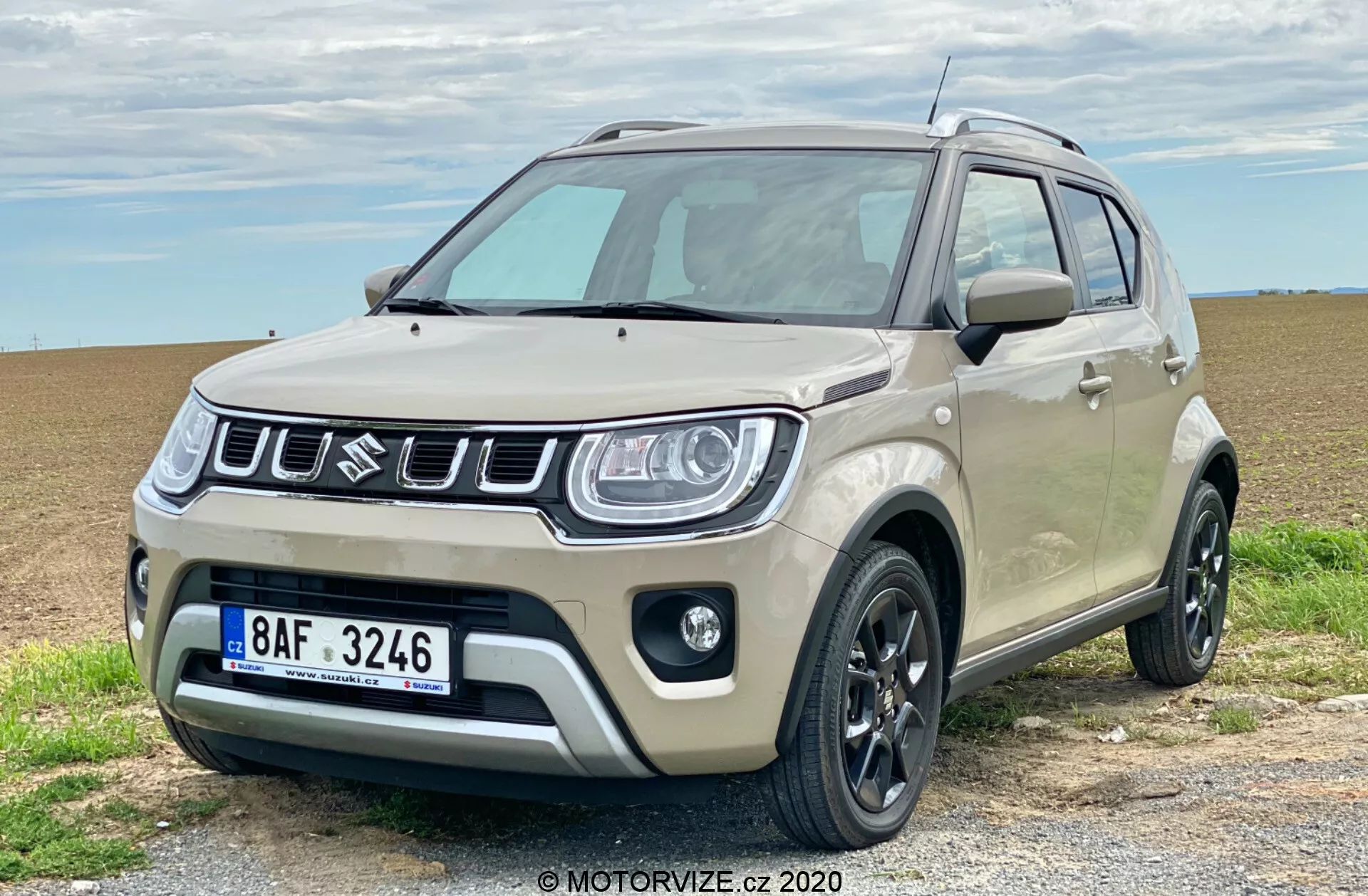 Kompaktowy SUV Suzuki Ignis 1.2 Dualjet Hybrid Club 2020 zaparkowany jest na żwirowej drodze z otwartym polem w tle, pokazując widok z przodu pod kątem 3/4. Ma dwukolorowe nadwozie z jasnobeżowym kolorem głównym i czarnymi akcentami na lusterkach bocznych, relingach dachowych i zderzakach. Charakterystyczny przedni grill ma pionowe listwy z centralnie umieszczonym logo marki. Nowoczesne reflektory z LED-owymi światłami do jazdy dziennej, czarne felgi aluminiowe, plastikowe osłony, światła przeciwmgielne i wysoki prześwit to wyróżniające cechy. Pojazd emanuje zarówno miejskim urokiem, jak i lekkimi możliwościami terenowymi.