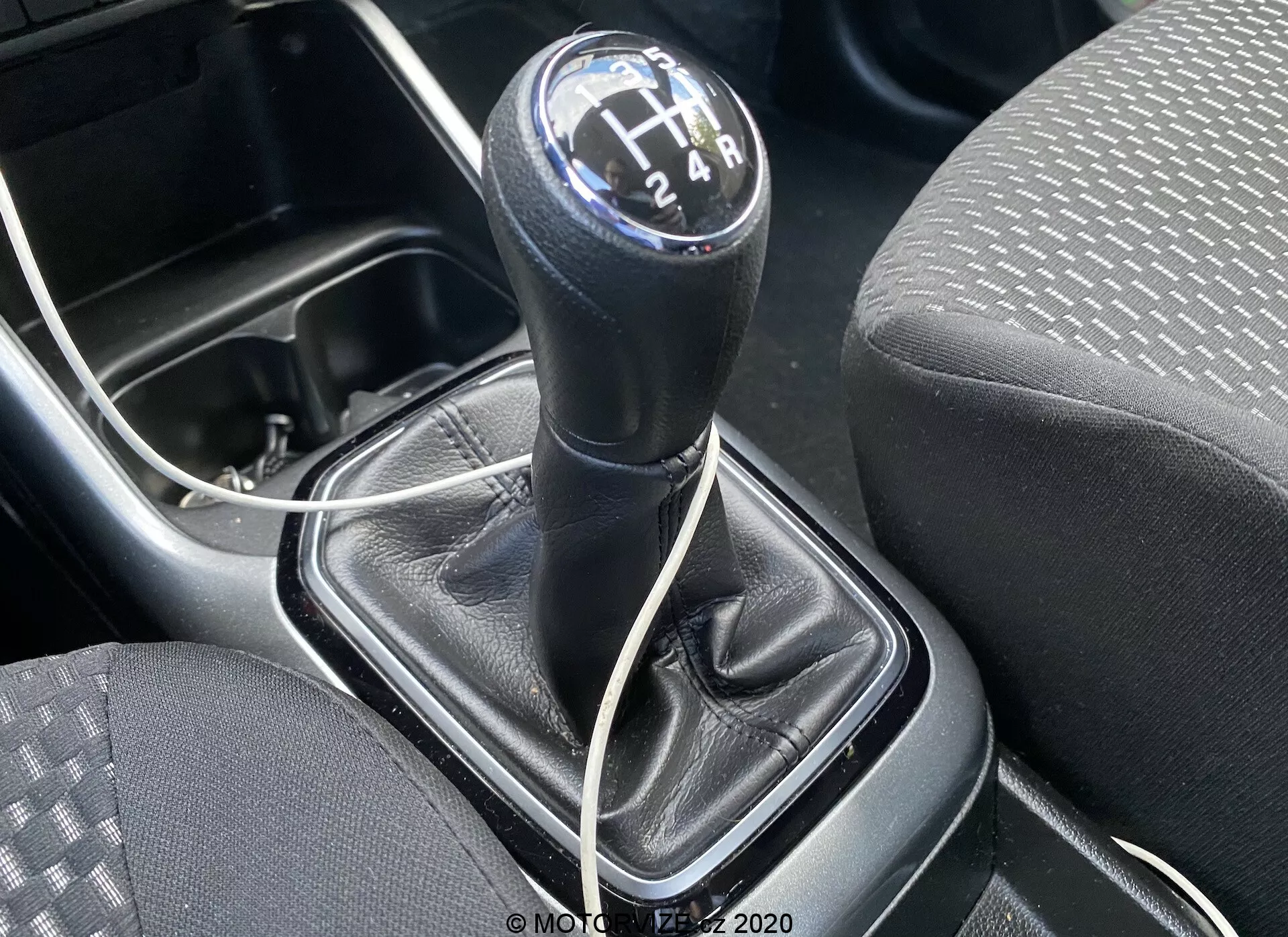 Zbliżenie na dźwignię zmiany biegów manualnej skrzyni biegów w Suzuki Ignis 1.2 Dualjet Hybrid Club 2020, wykonane z góry, skupiające się na okrągłym, czarnym gałce z białymi cyfry w układzie H z 6-przełożeniową skrzynią i biegiem wstecznym. Dźwignia zmiany biegów jest w pozycji neutralnej. Wokół dźwigni znajduje się czarna, skórzana osłona z widocznymi szwami oraz srebrna lub szara plastikowa konsola. Po lewej widoczna jest część siedzenia materiałowego z wzorem kropek i linii, a poniżej dźwigni widać biały kabel sugerujący obecność portu ładowania lub wejścia AUX. W dolnym lewym rogu widoczna jest krawędź uchwytu na kubek lub małego schowka, co podkreśla praktyczny design konsoli centralnej pojazdu.