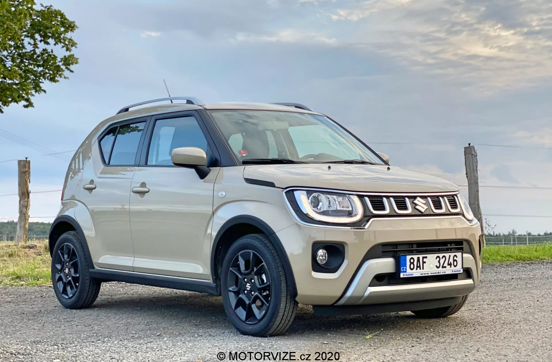 Obrazek przedstawia kompaktowe SUV Suzuki Ignis 2020 1.2 Dualjet Hybrid Club, zaparkowane na płaskiej, prawdopodobnie utwardzonej powierzchni, z częściowo pochmurnym niebem w tle. Uchwycony pod niskim kątem, przednia część pojazdu jest widoczna, podkreślając jego wysokość i prześwit. Samochód ma nowoczesny design z karoserią w jasnym kremowym lub beżowym kolorze i kontrastującym czarnym dachem. Posiada czarne, pięcioramienne alufelgi z solidnymi oponami, wąskie reflektory rozciągające się na błotniki oraz światła przeciwmgielne na dolnym zderzaku. Przedni grill prezentuje charakterystyczny design z logo Suzuki. Dwukolorowe boczne lusterka są dopasowane do karoserii i dachu, a na górze zamontowano relingi dachowe. Funkcje technologiczne mogą obejmować reflektory LED lub HID oraz światła do jazdy dziennej. Dolna część przedniego zderzaka ma wytrzymały design z symulowaną płytą ochronną. Widoczna jest również tablica rejestracyjna z przodu. Samochód zajmuje znaczną część kadru, podkreślając jego design i cechy w naturalnym świetle dziennym.