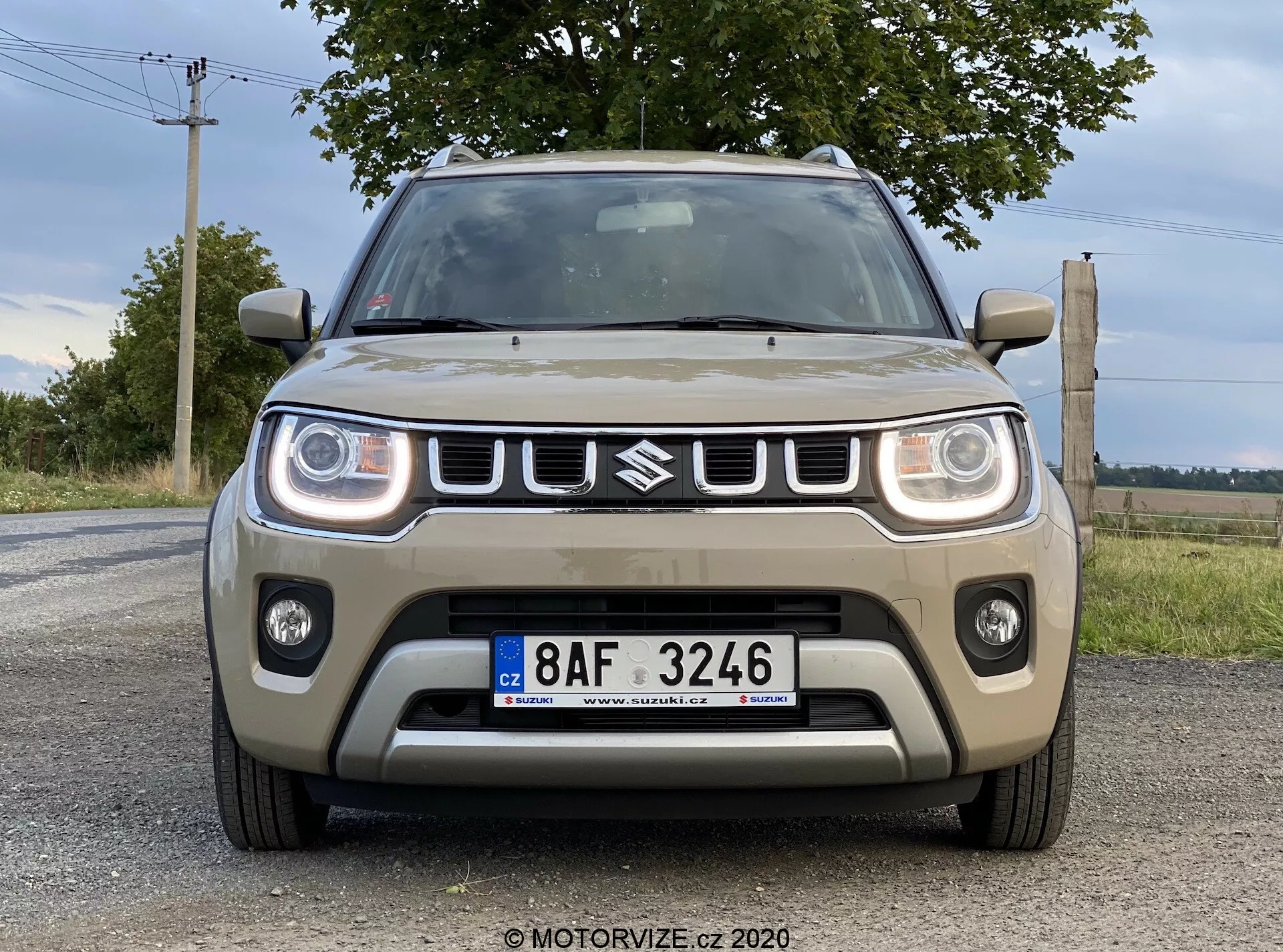 Widok z przodu Suzuki Ignis 2020 (Suzuki Ignis 1.2 Dualjet Hybrid Club) zrobiony z lekko niskiego kąta, ukazujący przednie cechy, w tym kratkę z emblematem, prostokątne reflektory LED, światła przeciwmgielne i centralny wlot powietrza na brukowanej drodze z naturalnym krajobrazem i zachmurzone niebo w tle.