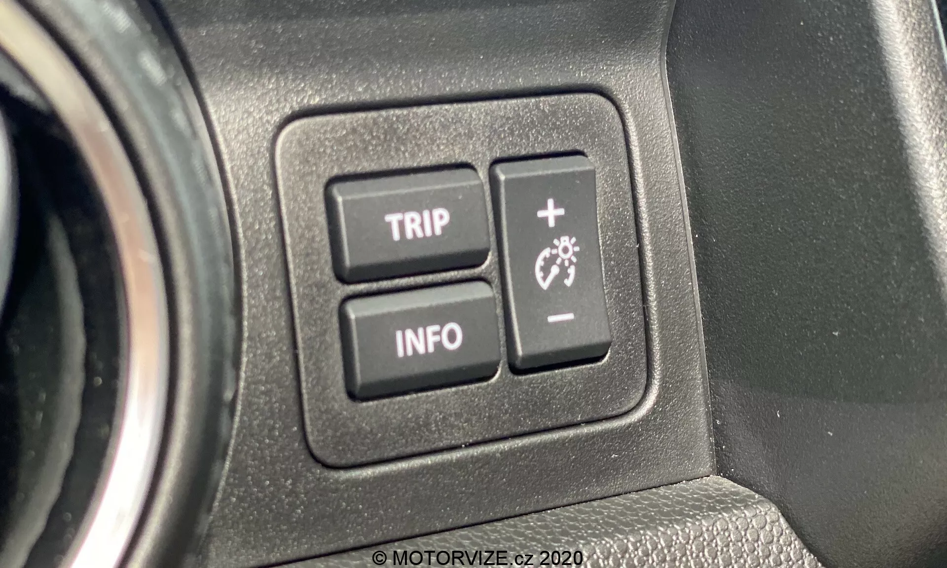 Zbliżenie na elementy sterujące wnętrza Suzuki Ignis 2020. Obraz skupia się na dwóch przyciskach oznaczonych jako 'TRIP' i 'INFO', które są częścią interfejsu komputera pokładowego pojazdu, umieszczone na teksturowanym panelu z tworzywa sztucznego. Przycisk 'TRIP' prawdopodobnie umożliwia dostęp do funkcji komputera podróży, podczas gdy przycisk 'INFO' wyświetla różne elementy informacyjne dotyczące samochodu. Zbliżenie podkreśla ergonomiczną konstrukcję i przyjazny dla użytkownika interfejs, podkreślając nowoczesną integrację technologiczną w pojeździe.