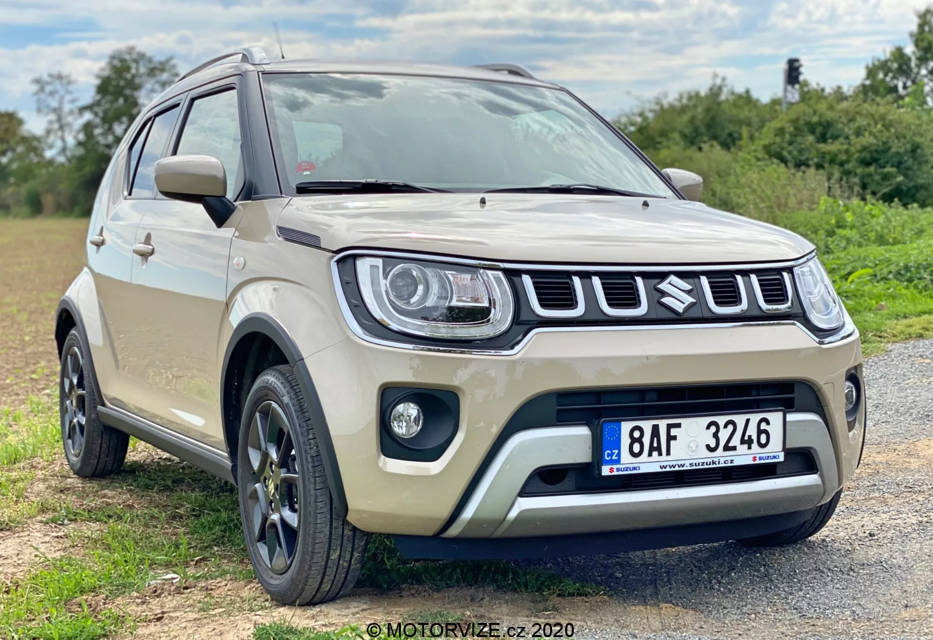 Nowoczesny subkompaktowy crossover SUV w kolorze beżowym, prawdopodobnie model Suzuki Ignis 2020, zaparkowany na szutrowej nawierzchni w okolicy półwieśniackiej. Pojazd, uchwycony z dolnego kąta w przednich trzech czwartych widoku, prezentuje swoją charakterystyczną przednią część z wyróżniającym się grillem, nowoczesnymi reflektorami i światłami przeciwmgielnymi. Wieloramienne felgi aluminiowe, zwiększony prześwit, ochronne obudowy oraz lusterka boczne z wbudowanymi kierunkowskazami podkreślają jego nowoczesne i solidne cechy. Mała głębia ostrości przy naturalnym oświetleniu skupia uwagę na samochodzie.
