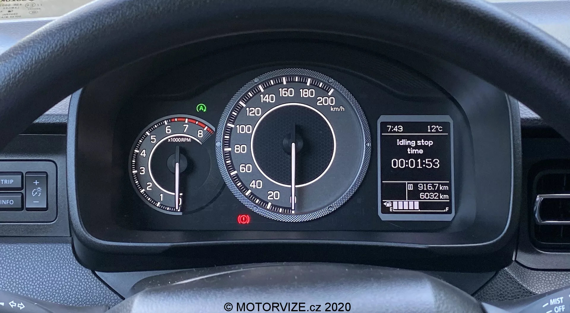 Zdjęcie wnętrza Suzuki Ignis 2020 skupione na zestawie wskaźników za kierownicą. Widoczna jest górna część kierownicy i dźwignie do kierunkowskazów i wycieraczek. Zestaw wskaźników łączy wyświetlacze analogowe i cyfrowe: po lewej analogowy obrotomierz z czerwonym polem przy 6500 obr./min i po prawej analogowy prędkościomierz do 220 km/h. Centralny wyświetlacz cyfrowy pokazuje temperaturę zewnętrzną (12°C), godzinę (7:43), funkcję 'Idling stop', stoper (00:01:53) i licznik kilometrów (91672 km). Nowoczesny, przyjazny dla użytkownika design typowy dla rynków metrycznych.