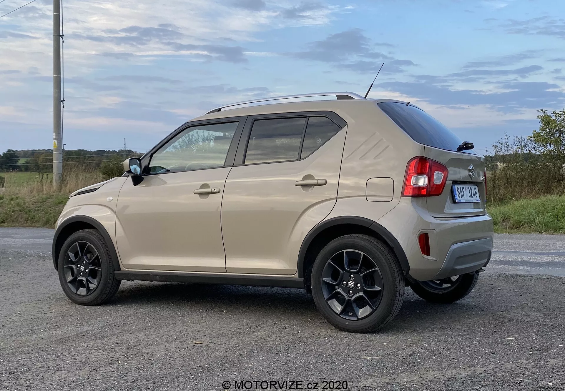Kompaktowy SUV Suzuki Ignis 1.2 DualJet Hybrid Club 2020 w widoku trzy-czwarte tyłu, z tylnym kapeluszem z wycieraczką, aerodynamiczną linią dachu, wysoko umieszczonym światłem stopu i anteną w kształcie płetwy rekina. Bok pojazdu pokazuje czarne wieloramienne obręcze ze stopów metali lekkich, kolorowe klamki drzwi z wbudowanymi kierunkowskazami, plastikowe osłony na dole drzwi i progach bocznych oraz czarne listwy wokół okien. Pojazd stoi zaparkowany w naturalnym wiejskim otoczeniu na utwardzonej powierzchni w ciągu dnia.