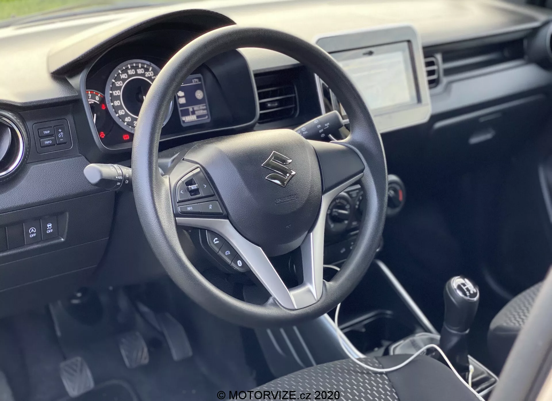 Wnętrze Suzuki Ignis 1.2 Dualjet Hybrid Club 2020 z trójramienną kierownicą z wbudowanymi sterownikami do audio, połączeń bezrękawiczkowych i tempomatu. Deska rozdzielcza zawiera analogowe wskaźniki, centralny ekran dotykowy systemu informacyjno-rozrywkowego oraz dźwignię zmiany biegów manualnych. Praktyczne wnętrze w dwóch odcieniach kładzie nacisk na funkcjonalność z trwałymi materiałami i dobrą widocznością w naturalnym świetle.