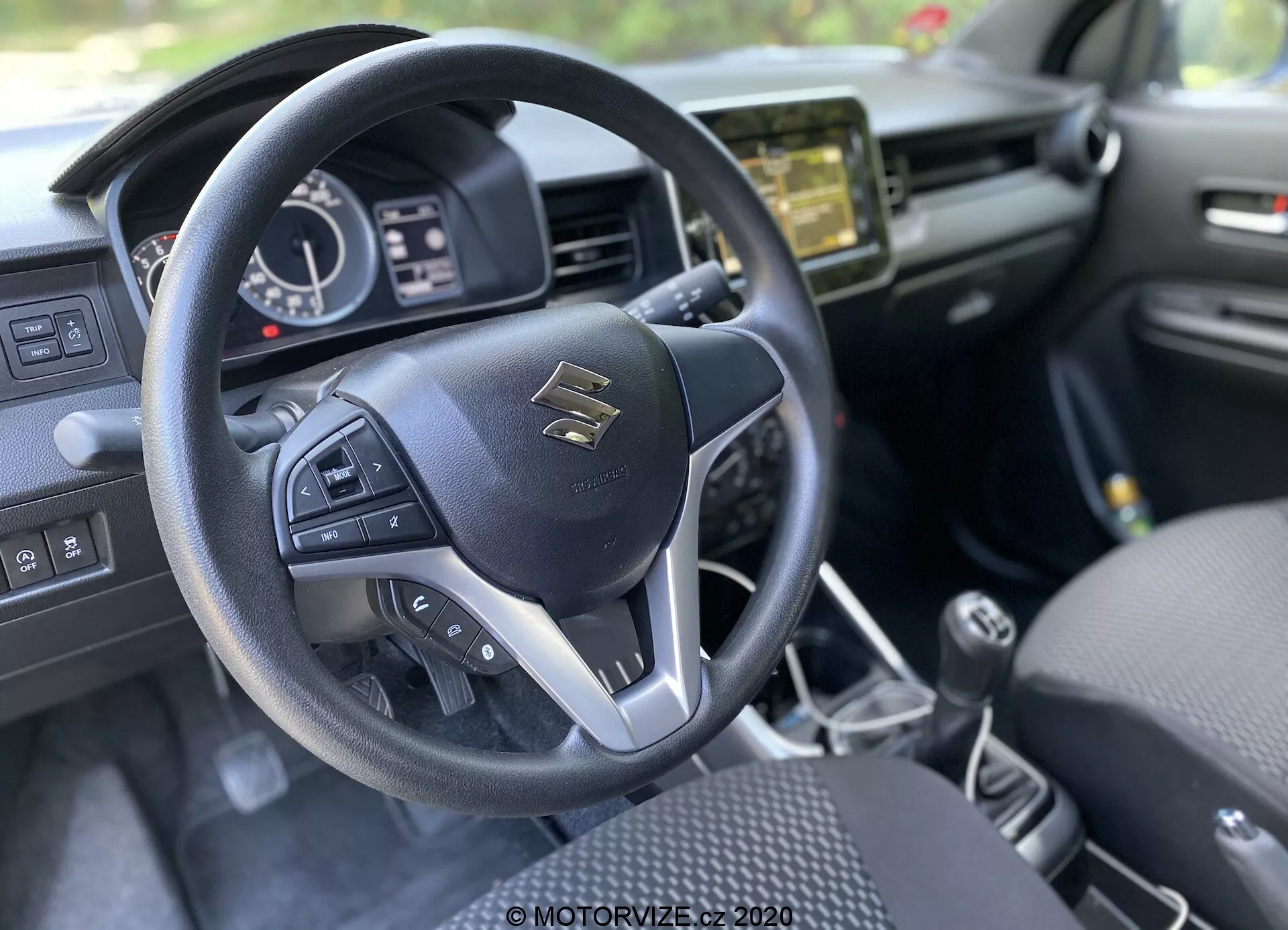 Widok wnętrza z perspektywy kierowcy Suzuki Ignis 2020 (1.2 Dualjet Hybrid Club) koncentrujący się na nowoczesnej, trójramiennej kierownicy z przyciskami wielofunkcyjnymi. Znak Suzuki znajduje się na środku, otoczony srebrnymi akcentami i jakością przeszyć. Za kierownicą częściowo widoczny zestaw wskaźników z cyfrowym wyświetlaczem i analogowymi tarczami pokazuje prędkość, obroty oraz stan pojazdu. Na centralnej desce rozdzielczej znajduje się ekran systemu multimedialnego, prawdopodobnie oferujący audio, nawigację i ustawienia pojazdu. Konsola centralna ma manualną skrzynię biegów, sugerując manualną skrzynię. Wystrój wnętrza jest głównie czarny z teksturowymi elementami, siedzenia z wzorzystym wzorem, co sugeruje estetykę i komfort.