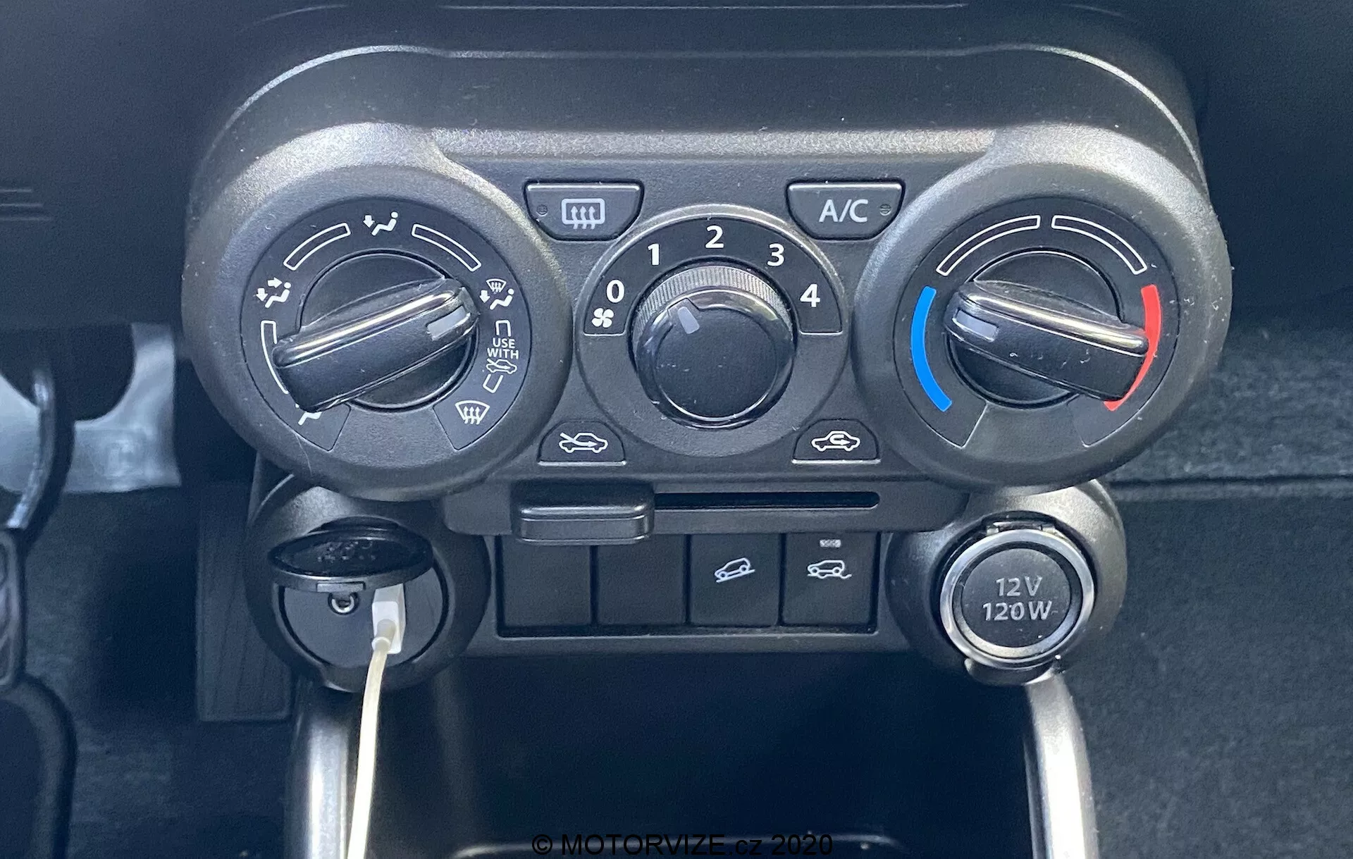 Zbliżenie konsoli środkowej Suzuki Ignis 2020, koncentrujące się na jednostce sterowania klimatyzacją z trzema głównymi pokrętłami na temperaturę, prędkość wentylatora i kierunek przepływu powietrza. Pokrętło temperatury ma niebieskie i czerwone wskaźniki dla chłodniejszych i cieplejszych ustawień. Nad centralnym pokrętłem prędkości wentylatora znajduje się przycisk 'A/C', przycisk cyrkulacji powietrza w kabinie i przyciski odmrażania przedniego. Pod kontrolkami znajduje się gniazdo 12V 120W, podkreślające praktyczny i przyjazny dla użytkownika design pojazdu z ręcznymi przełącznikami.