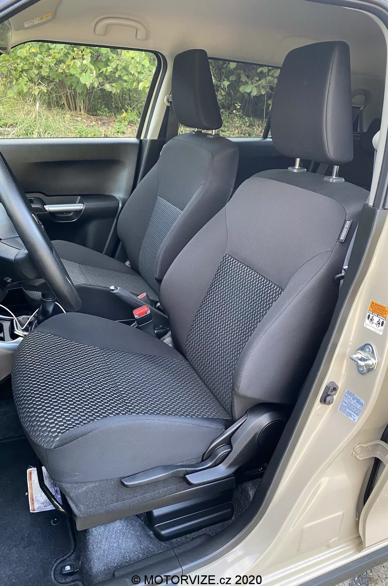 Profesjonalne zdjęcie wnętrza Suzuki Ignis 2020 (Suzuki Ignis 1.2 Dualjet Hybrid Club), skupiające się na obszarze siedzenia przedniego pasażera. Wykonane z wysokiej perspektywy, aparat uchwycił przejrzysty widok z otwartymi drzwiami samochodu. Tapicerka z tkaniny dwukolorowej na siedzeniu ma wzorzysty design dla trwałości. Regulacje siedzenia są manualne, z zagłówkiem, pasem bezpieczeństwa i widocznymi dźwigniami do regulacji. Widoczna jest również część konsoli środkowej i wewnętrzny panel drzwi z etykietami regulacyjnymi. Ciemna wykładzina i jasny próg drzwi podkreślają kontrast designu. Zdjęcie przypisane do MOTORVIZE.cz 2020.