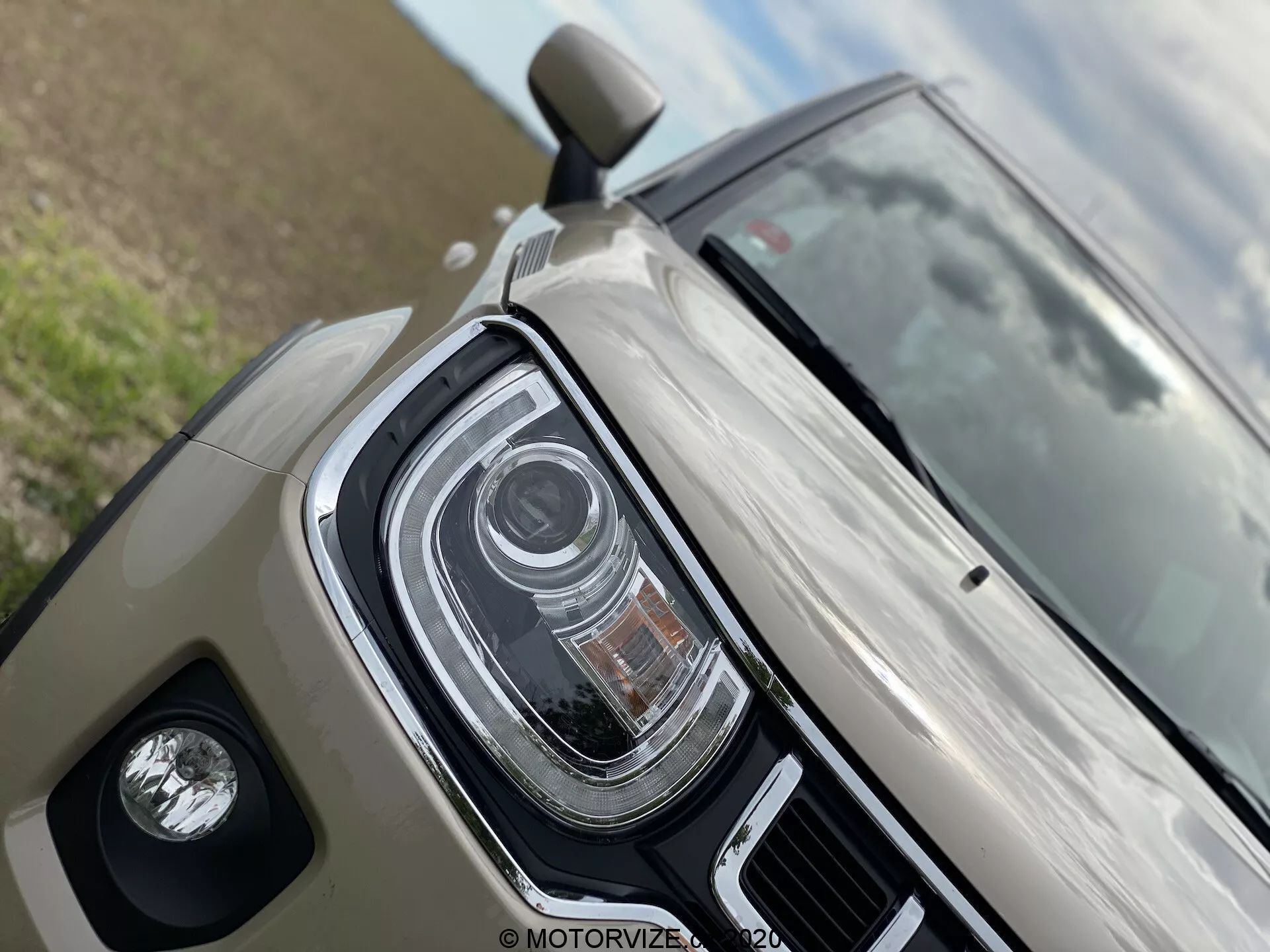 Zbliżenie na Suzuki Ignis 2020, ukazujące przedni lewy kwadrant. Charakteryzuje się konstrukcją reflektorów LED z wieloma przedziałami, prawdopodobnie adaptacyjnymi reflektorami lub automatycznymi światłami drogowymi. Pokazuje błyszczące wykończenie w odcieniu beżowym/jasnoszarym i obudowę świateł przeciwmgielnych. Częściowy widok lusterka bocznego sugerujący zaawansowaną technologię, taką jak wskaźniki w lusterkach bocznych lub monitorowanie martwego pola.