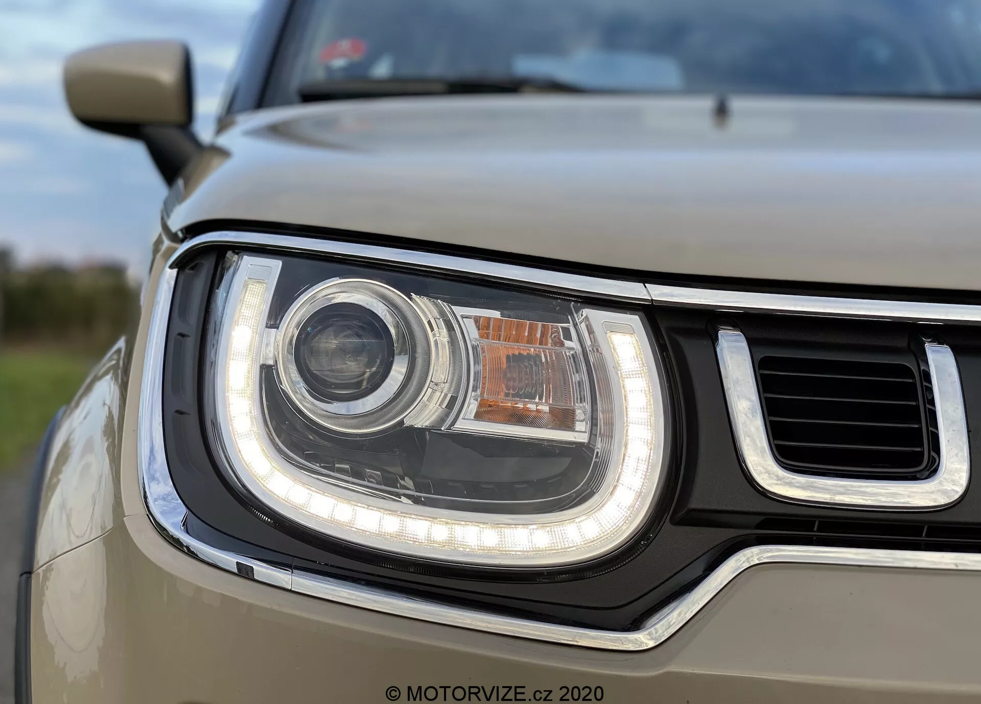 Zbliżenie zespołu reflektorów na przedniej lewej stronie Suzuki Ignis 2020, z widocznym okrągłym głównym reflektorem prawdopodobnie LED lub HID, otoczonym LED-owym światłem dziennym (DRL) w formie rury. Reflektor posiada mniejsze, pomarańczowe światło kierunkowskazu lub postojowe umieszczone powyżej. Częściowo widoczna kratka jest wyposażona w poziome pasy, a projekt reflektora ma zarówno zakrzywione, jak i proste krawędzie, co tworzy nowoczesny, solidny wygląd. Głębia ostrości skupia się na reflektorze, podczas gdy tło i inne części auta są lekko rozmazane.