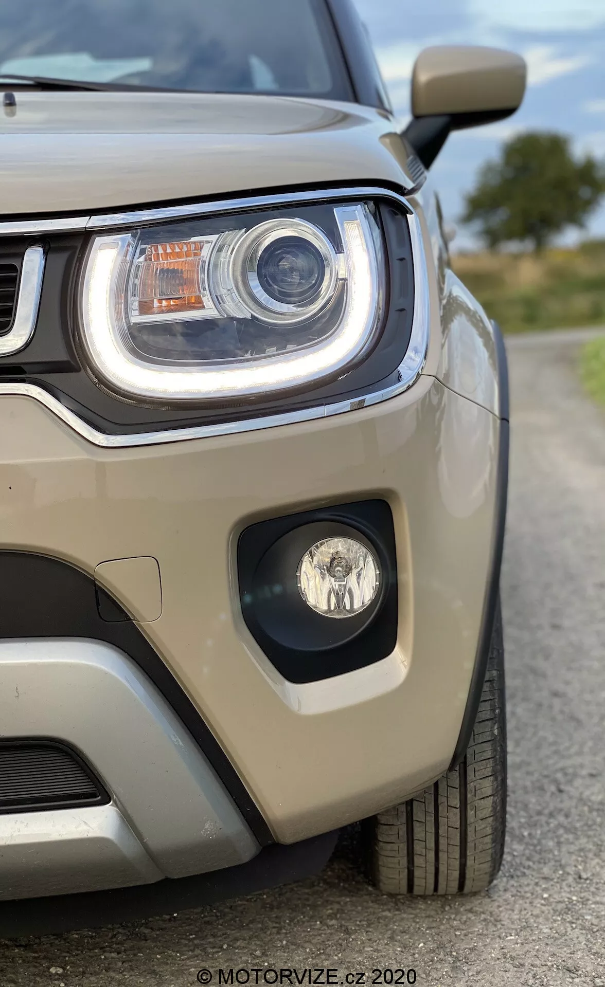 Zbliżony widok lewej strony przodu Suzuki Ignis 2020 z naciskiem na zespół reflektora i część grille. Obraz, wykonany pod nieco niskim kątem, podkreśla agresywne linie reflektora. Charakteryzuje się wyraźnym światłem do jazdy dziennej LED, technologią projektorową, dodatkowymi elementami oświetleniowymi i światłem przeciwmgielnym w osobnej oprawie. Jasnobeżowy metaliczny lakier odbija światło, a widoczny czujnik parkowania na zderzaku zwraca uwagę na technologię wspomagania parkowania. Auto wygląda na bardzo czyste i dobrze utrzymane, co przyciąga uwagę do jego nowoczesnego designu.