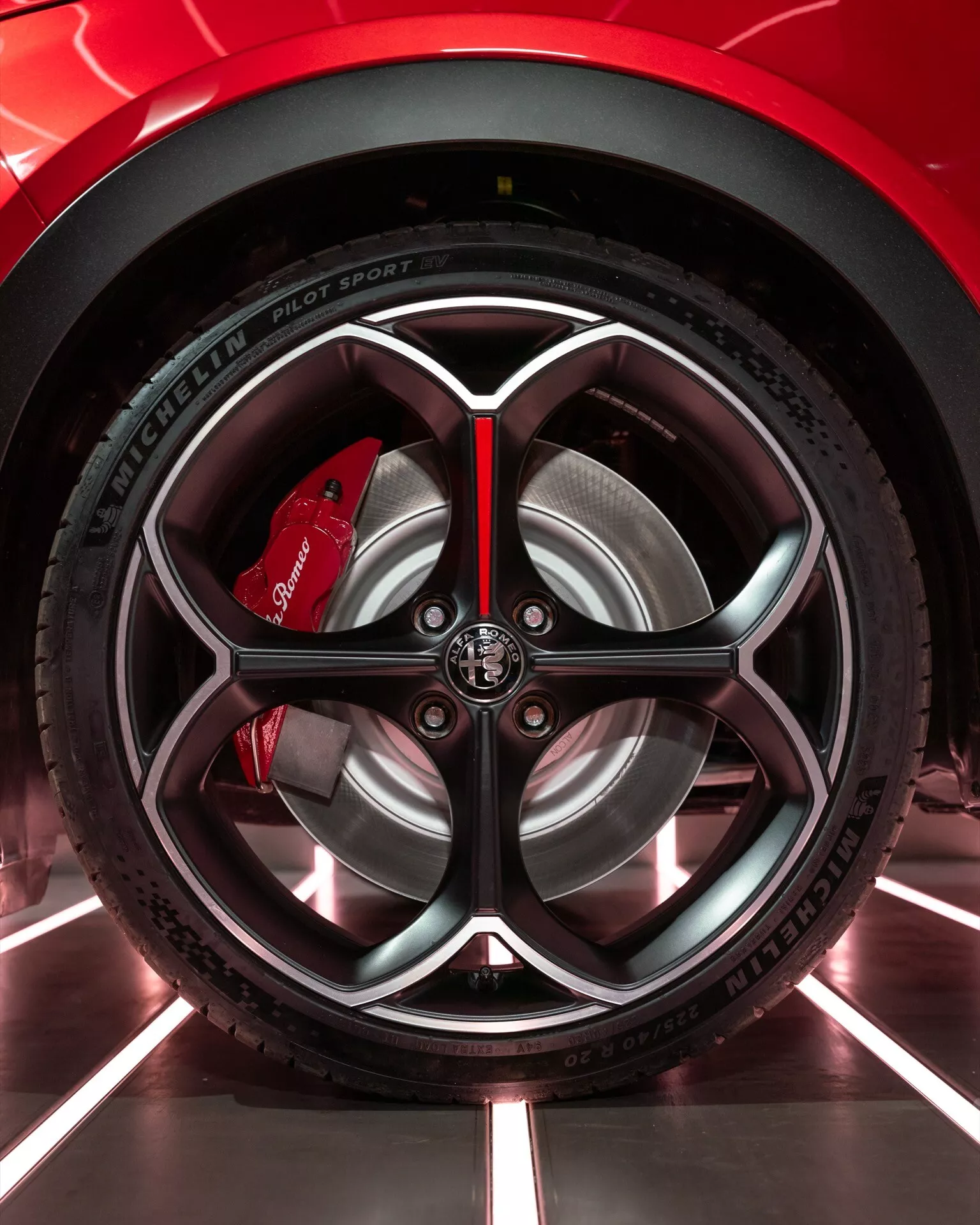 Szczegółowe zbliżenie na koło Alfa Romeo Junior 2024 pokazujące złożony design felgi i oponę Michelin Pilot Sport 4 w środowisku salonu, podkreślające wydajność i sportową estetykę