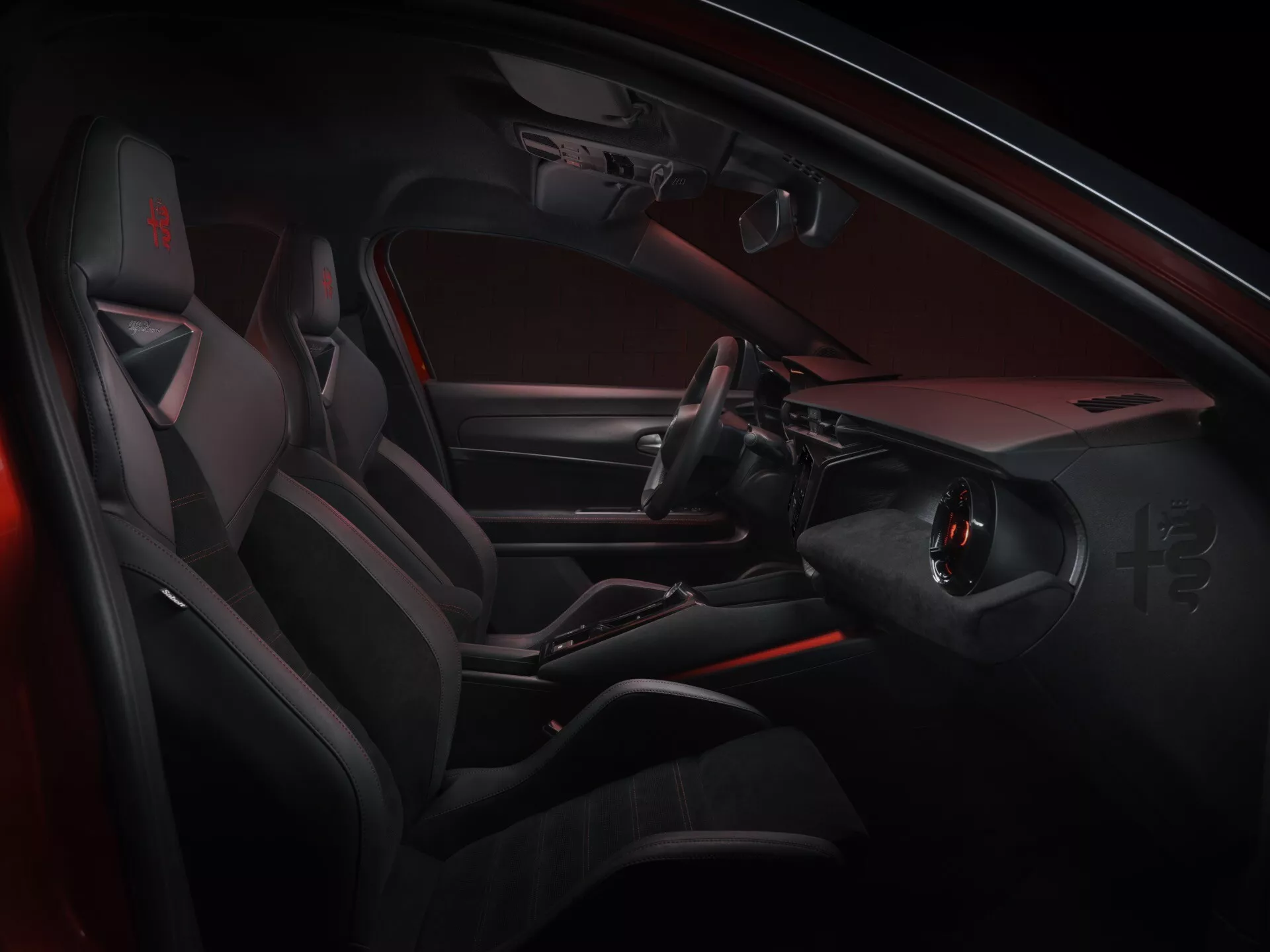 Widok wnętrza Alfa Romeo Junior 2024 pokazujący stronę kierowcy z nowoczesnymi, sportowymi cechami projektu, w tym płaskie dno kierownicy z zintegrowanymi przyciskami, elegancka konsola środkowa bez tradycyjnej dźwigni zmiany biegów, sportowe siedzenia z wyraźnymi bocznymi wzmocnieniami i luksusowe materiały z akcentami czerwonego oświetlenia ambientowego