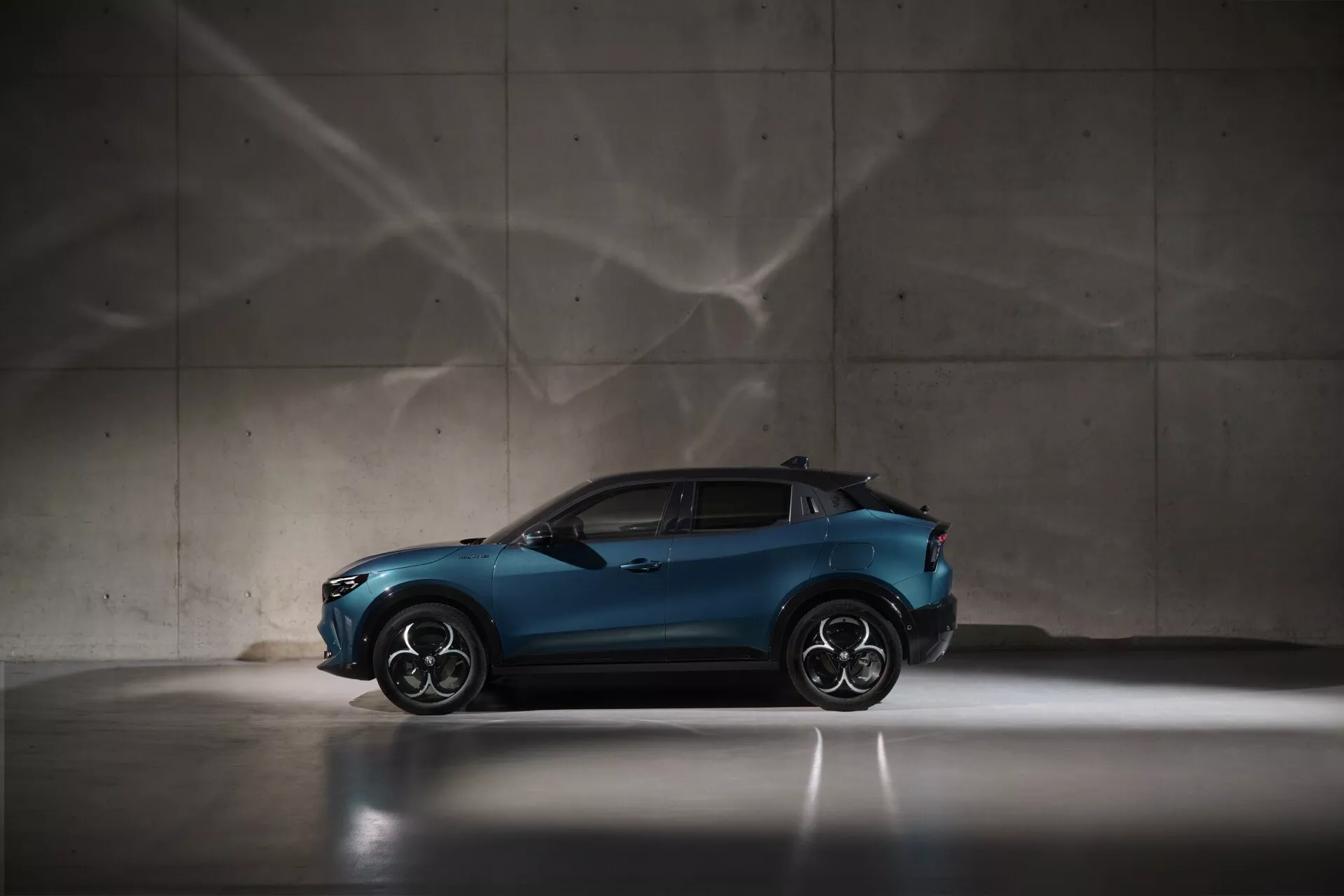 Alfa Romeo Junior 2024 w metalicznym niebieskim kolorze, widok z boku podkreślający sportowy i kompaktowy design crossovera, z dużymi felgami ze stopu i opadającą linią dachu, dramatyczne oświetlenie i artystyczne wzory cieni w tle