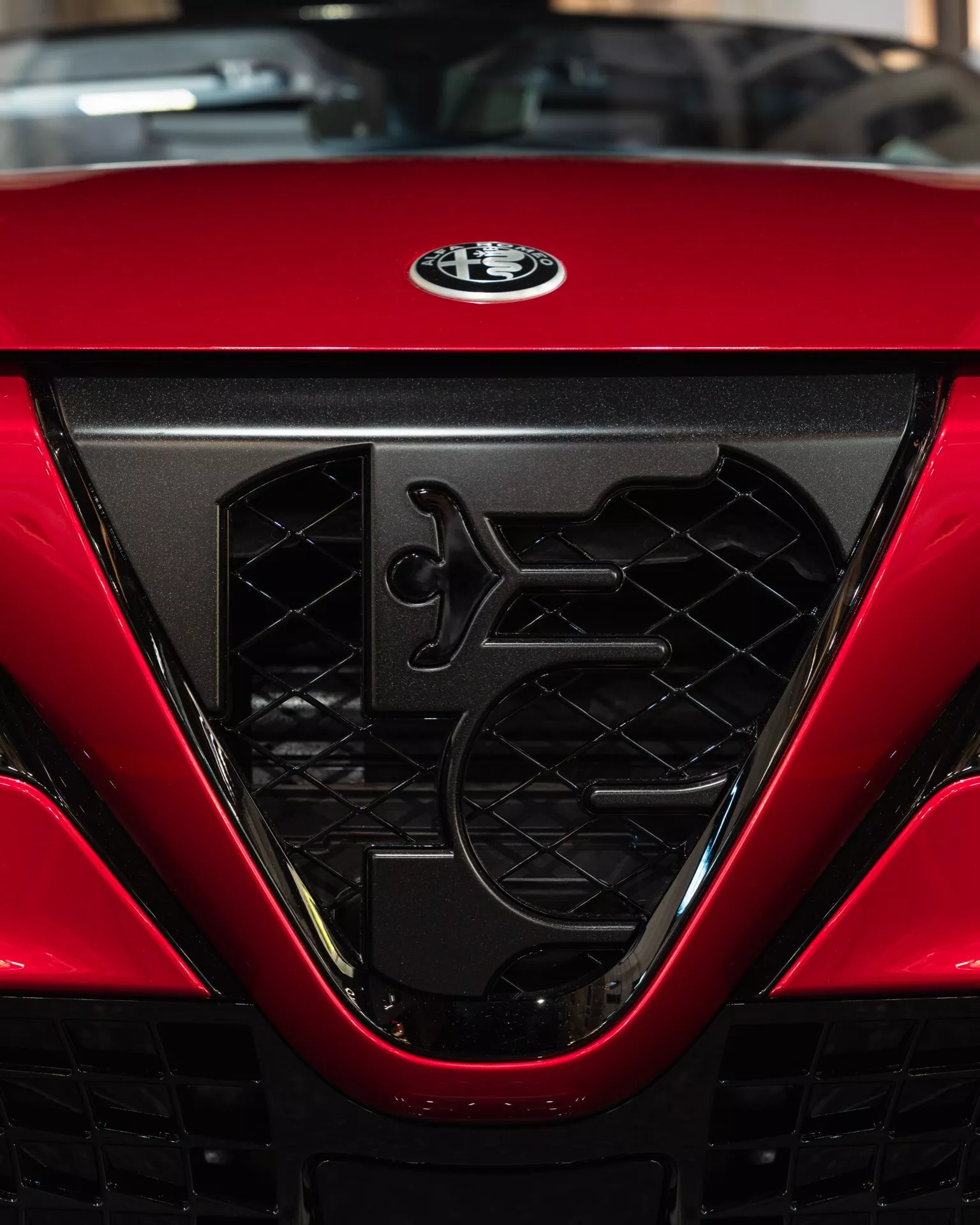 Zbliżenie na grill i emblem producenta Alfa-Romeo Junior (966) 2024, podkreślający złożony heksagonalny design siatki, cechy aerodynamiczne i błyszczące czerwone wykończenie
