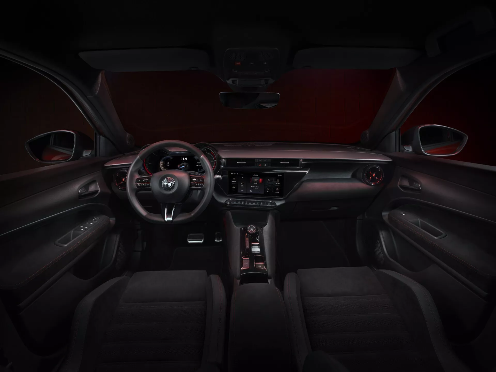 Widok wnętrza Alfa-Romeo Junior 2024 (966) prezentujący tapicerkę z ciemnej skóry z detalami szycia na przednich siedzeniach, kierownicę z trzema ramionami z zamontowanymi sterowaniami zawierającymi emblemat producenta, częściowo widoczny cyfrowy zestaw wskaźników, centralny system rozrywki z ekranem dotykowym z przyciskami sterowania klimatyzacją i innymi funkcjami samochodu poniżej, elegancka dźwignia zmiany biegów obok intuicyjnego pokrętła sterującego, podkreślone kontrastującą czerwoną wyściółką dachu i panelami drzwi z wbudowanymi sterowaniami i detalami szycia.
