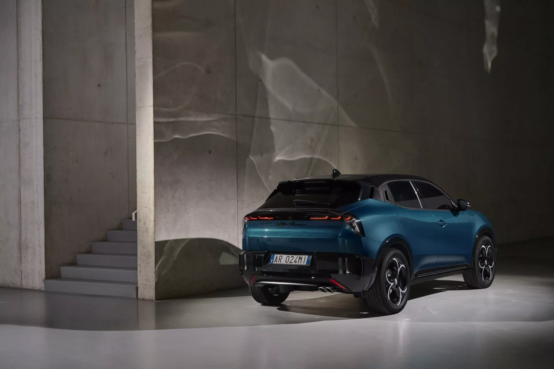 Alfa Romeo Junior 2024 widziany z tyłu z perspektywy trzech czwartych prezentuje nowoczesny design, pełnej długości światła tylne, sportową sylwetkę przypominającą coupe z czterema drzwiami oraz luksusowe elementy, wystawiony w eleganckim wnętrzu z odbijającą podłogą i betonowymi konstrukcjami.