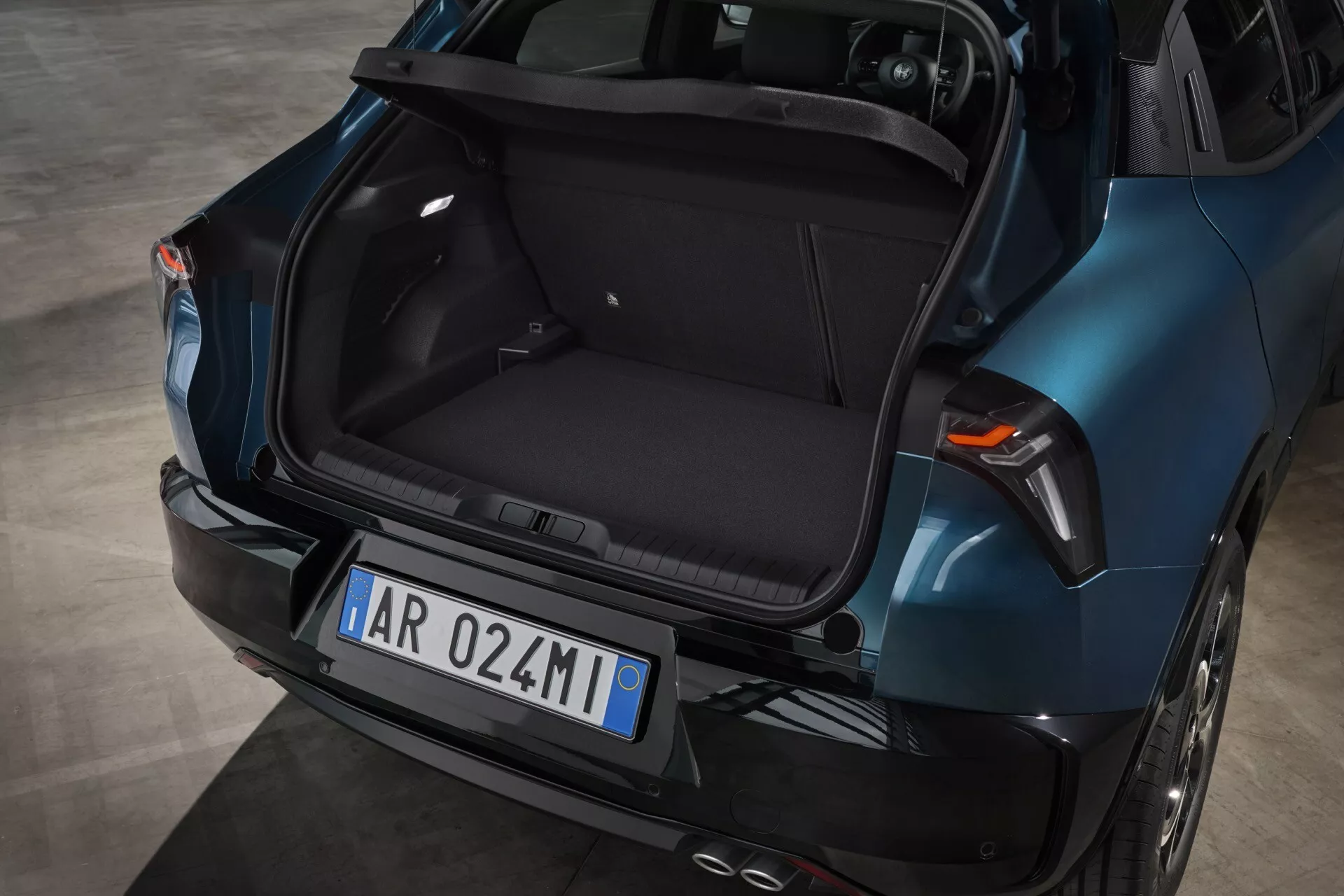 Tylny widok Alfa Romeo Junior 2024 ukazujący pusty bagażnik, metaliczny niebieski lakier, nowoczesny design z aerodynamicznymi liniami, zintegrowane światła hamowania w klapie bagażnika, tylne światła LED i podwójny układ wydechowy.