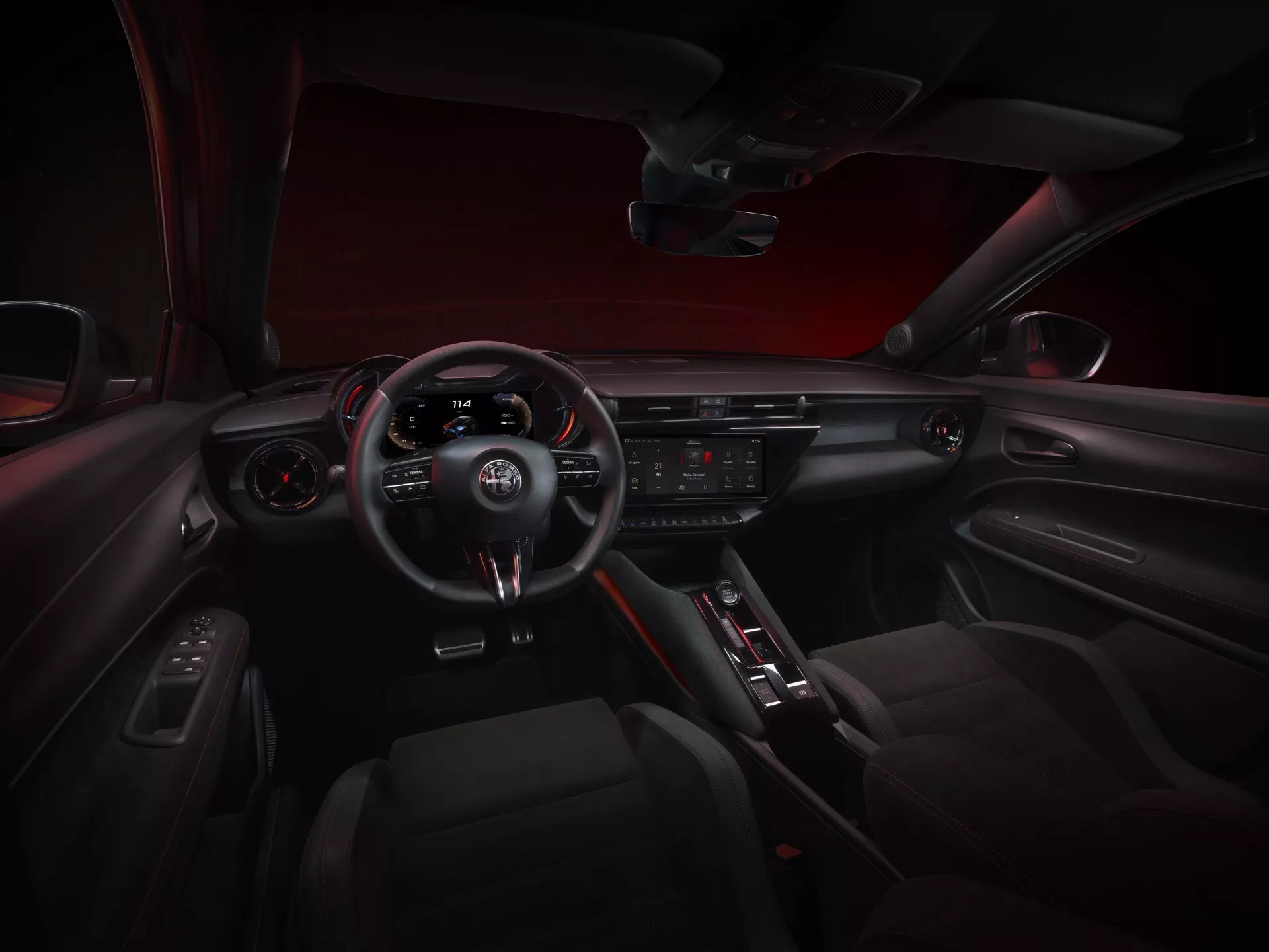 Widok wnętrza kokpitu Alfa-Romeo Junior 2024 z perspektywy centralnego tylengo siedzenia, przedstawiający deskę rozdzielczą, sportową kierownicę z trzema ramionami z zintegrowanymi sterowaniami i łopatkami zmiany biegów, panel przyrządów, centralny wyświetlacz systemu infotainment, przyciski sterowania klimatyzacją, selektor automatycznej skrzyni biegów, panel drzwi z kontrolami elektrycznymi i sportowo-elegancką atmosferę z czerwonym oświetleniem akcentującym