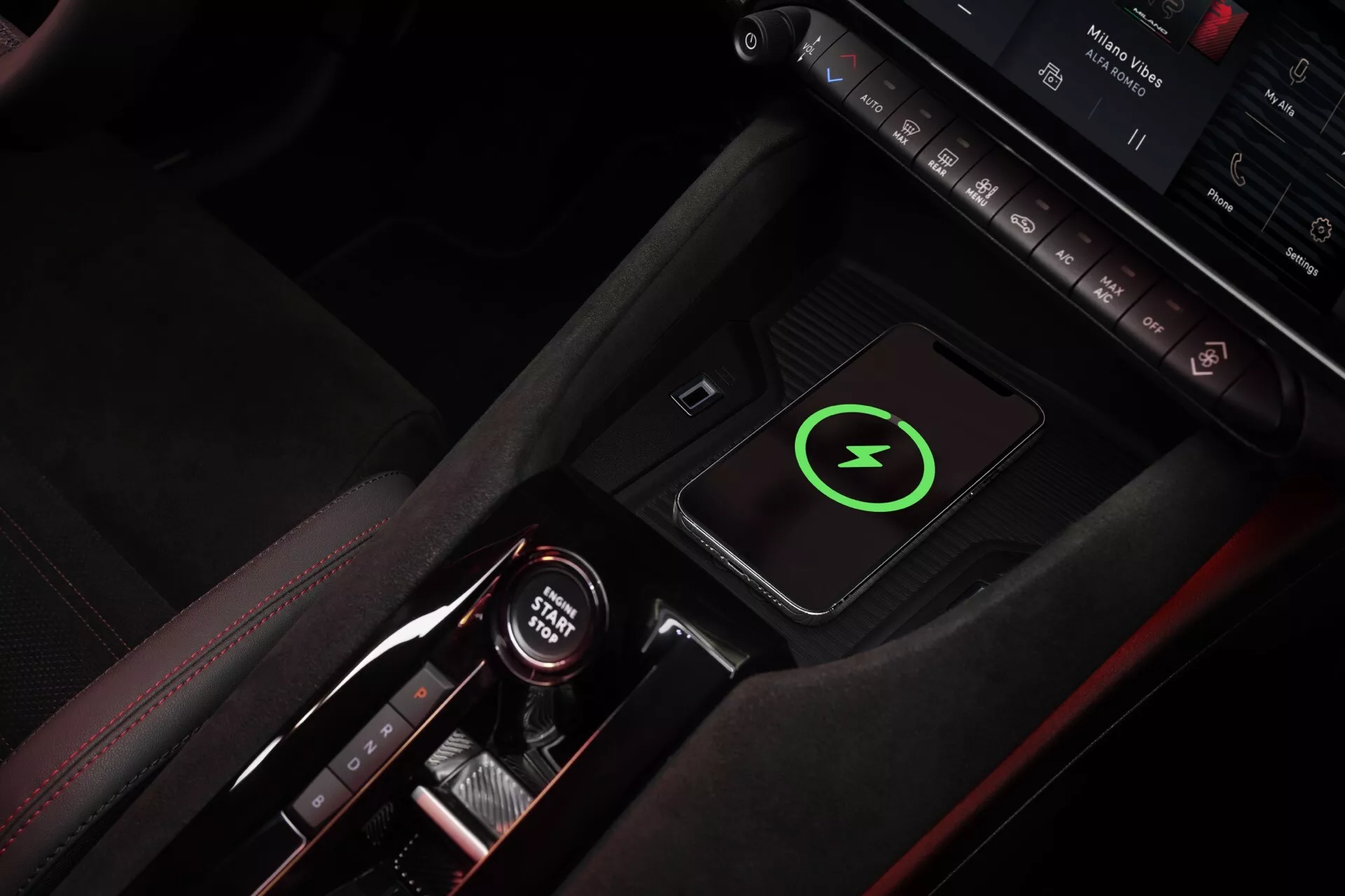 Widok z góry na konsolę środkową Alfa-Romeo Junior 2024, podkreślający czerwone przeszycia, elegancki dźwignię zmiany biegów z wskaźnikami automatycznej skrzyni biegów, bezprzewodową podkładkę do ładowania z zieloną ikoną błyskawicy, przyciski sterowania, ambientowe oświetlenie, uchwyt na kubek z czerwonymi akcentami, akcentujący luksusowy i technologicznie zorientowany design wnętrza