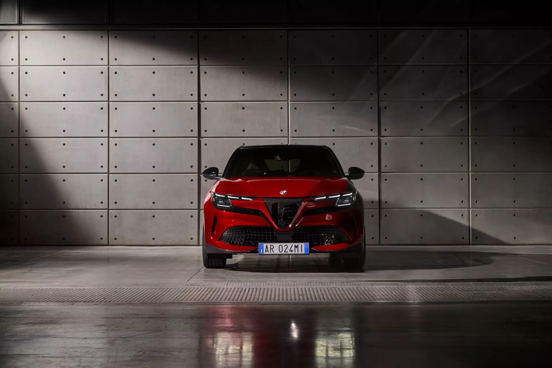 Przedni widok Alfa Romeo Junior 2024 uchwycony z niskiego kąta na metalowej kratownicowej podłodze, z LEDowymi reflektorami, wyraźną atrapą z emblematem producenta i eleganckim projektem zderzaka, na tle betonowej ściany z okrągłymi wgnieceniami, pod dramatycznym oświetleniem ambientalnym.