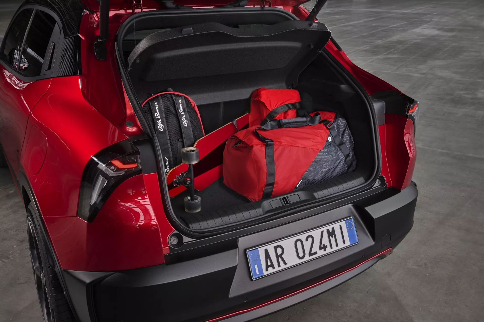 Czerwony model Alfa-Romeo Junior 2024 z otwartym bagażnikiem ukazującym przestronne wnętrze, fotelik samochodowy dla dziecka i czerwoną torbę podróżną, z sportowym tylnym designem i nowoczesnymi diodami LED, demonstrujący potencjał do przyjazności rodzinnej i technologii asysty parkingowej