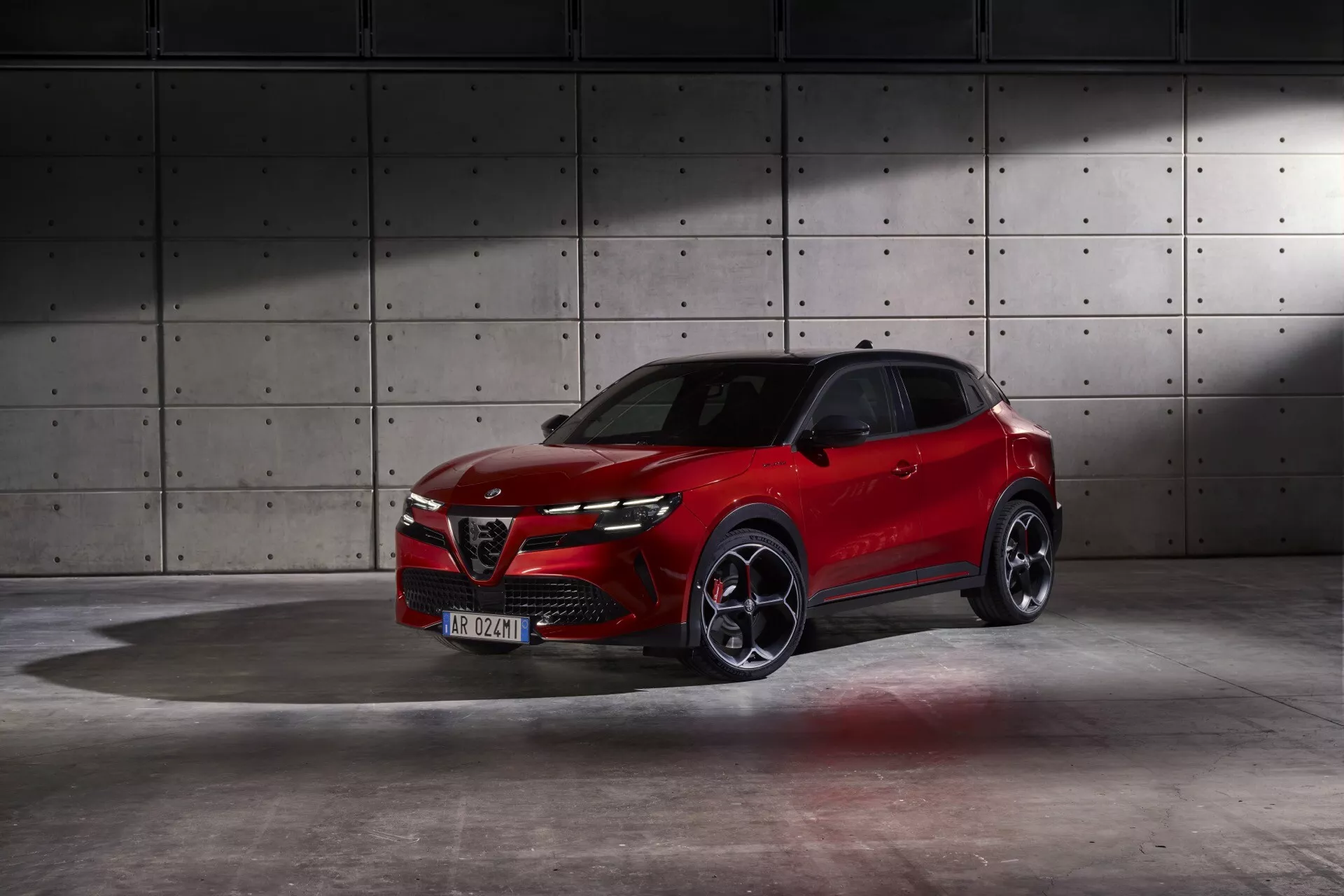 Alfa Romeo Junior 966 z 2024 roku w żywym czerwonym kolorze prezentujące dynamiczny design i zaawansowane funkcje w profesjonalnym studiu z industrialnym tłem